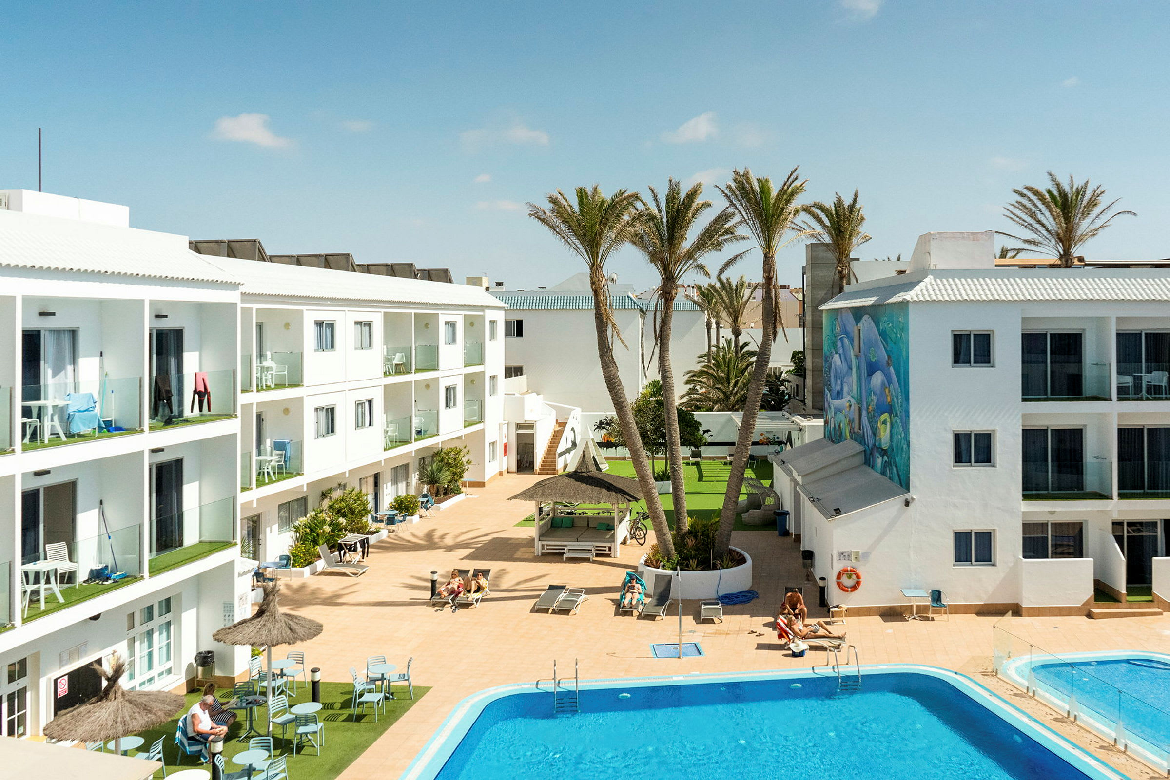 Hotellområde med vita byggnader, en pool och solstolar. Palmer och en dekorerad vägg är synliga. (AI-genererad text)