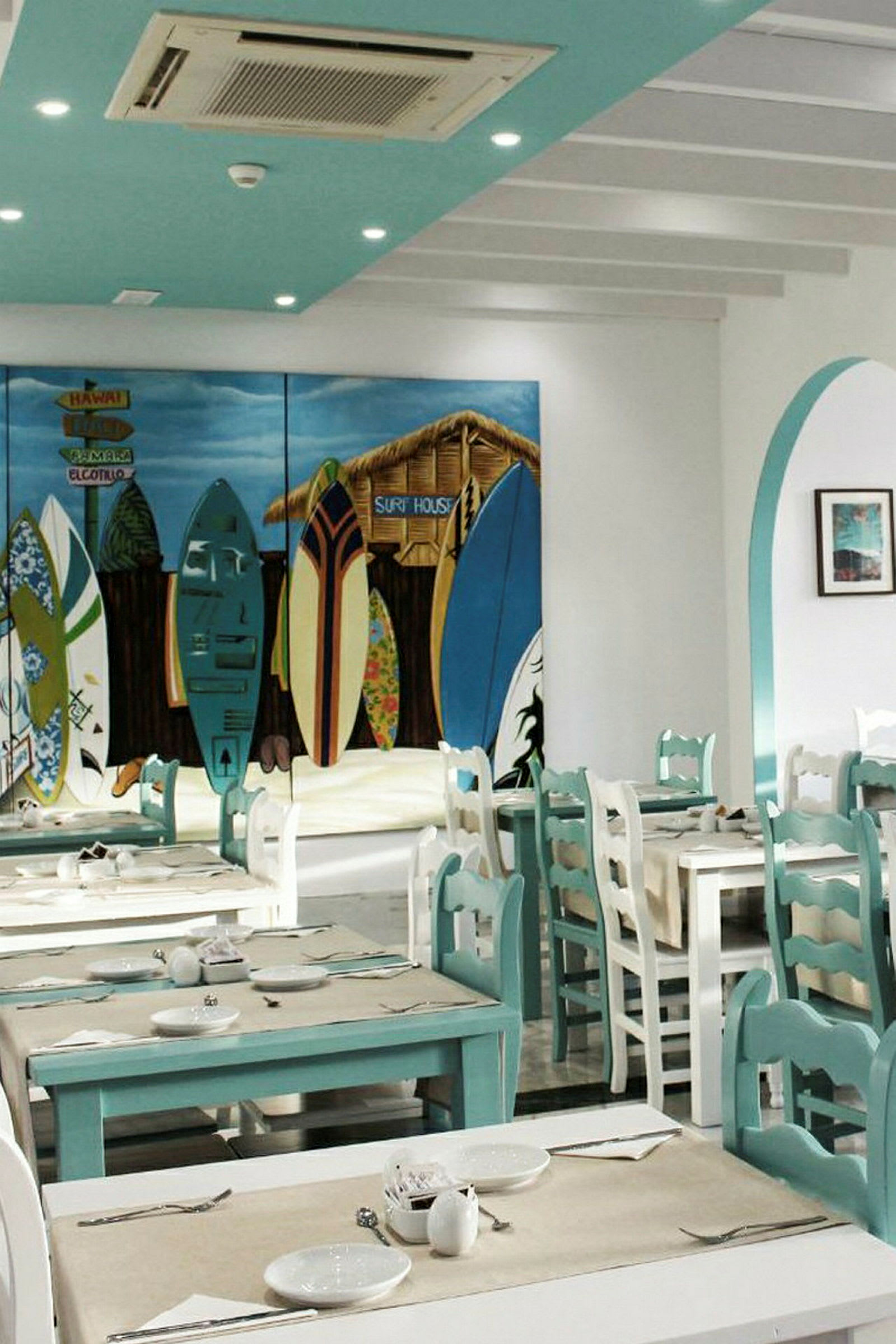 En interiör av en restaurang med turkosa och vita bord och stolar. En färgglad väggmålning visar surfbrädor och en hydda. (AI-genererad text)