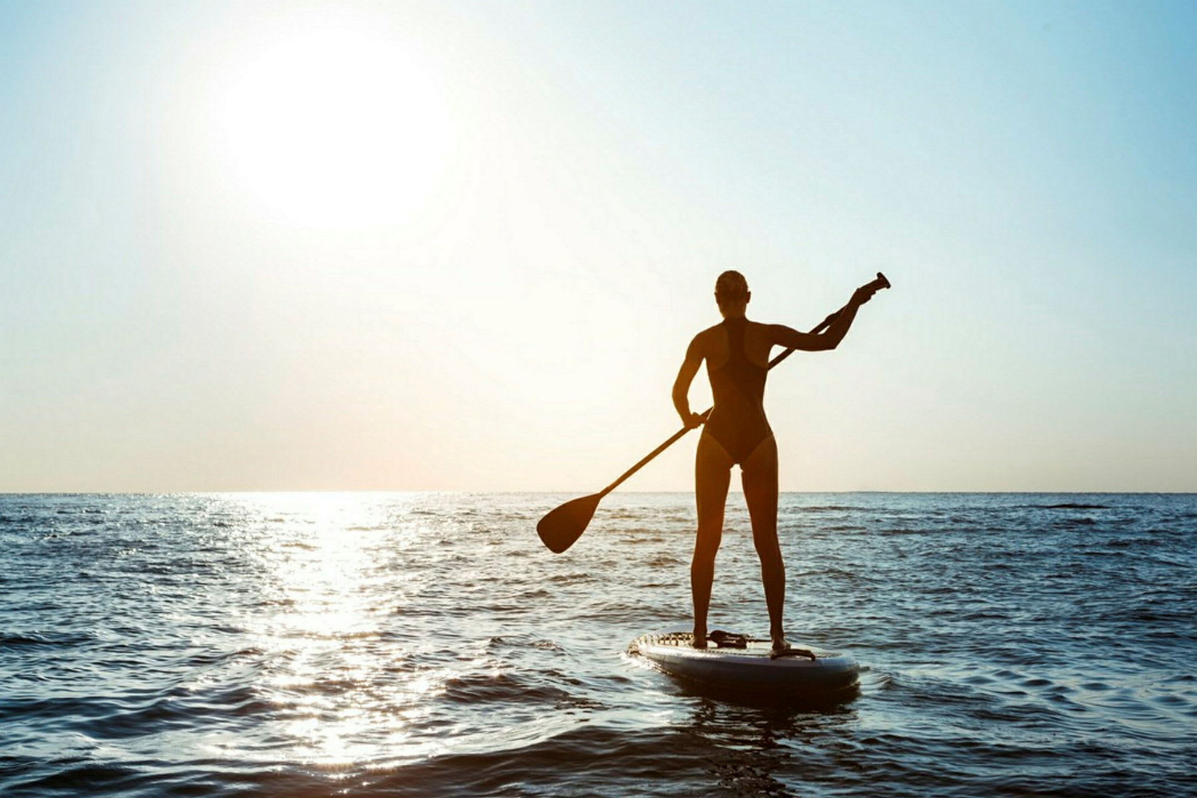En person paddlar stående på en paddleboard på havet i motljus med solen i bakgrunden. (AI-genererad text)