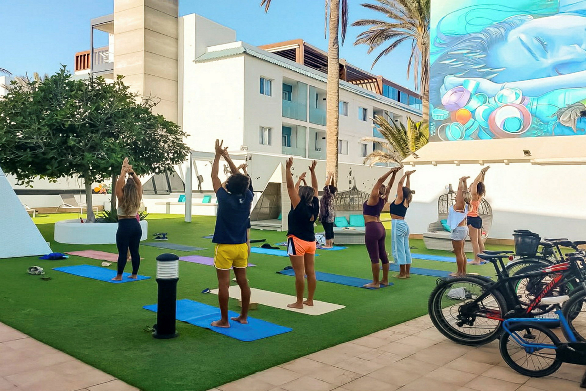 En grupp människor utför yoga utomhus på gräs med mattor, omgivna av en modern byggnad och palmer. (AI-genererad text)