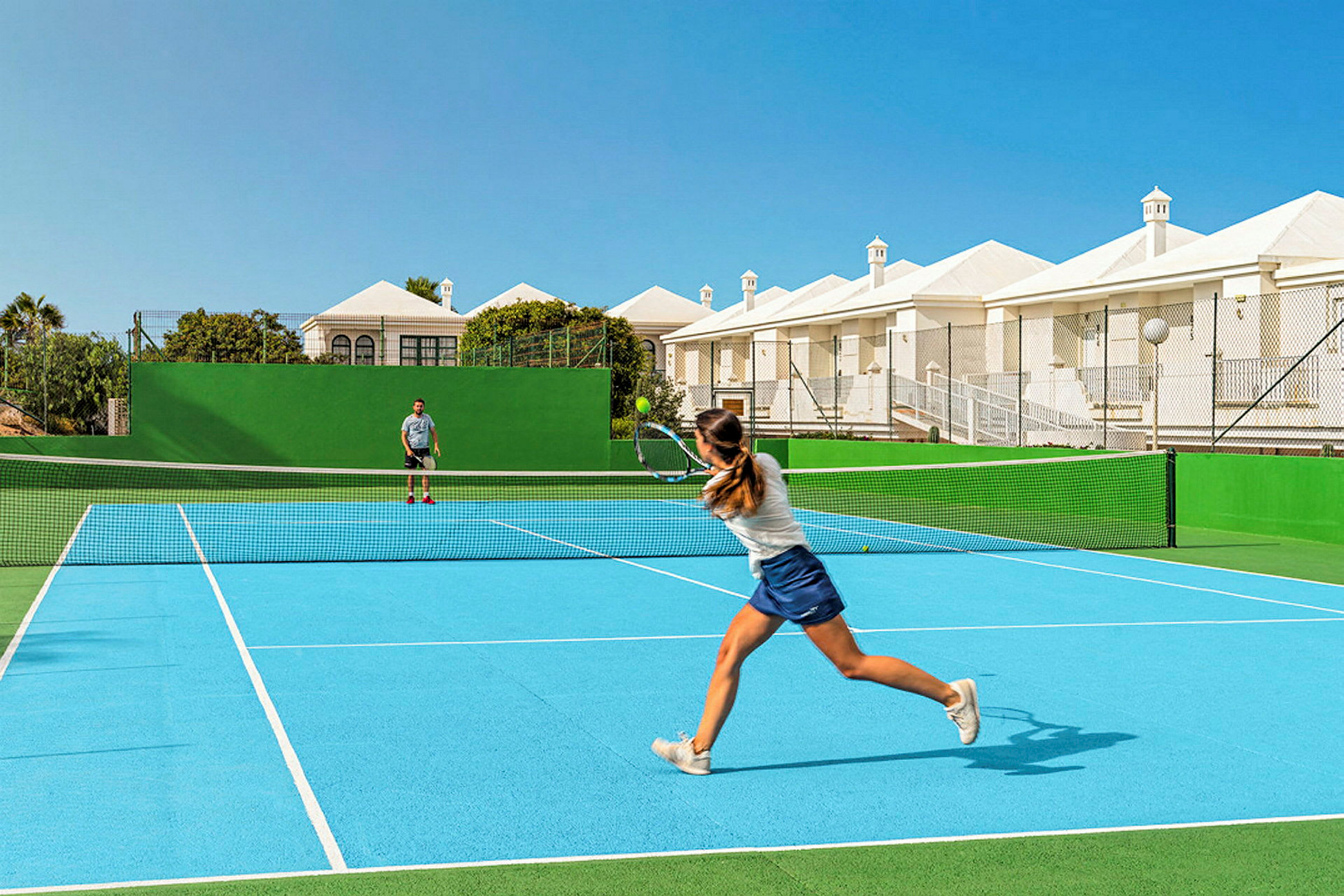 Två personer spelar tennis på en utomhus tennisbana med blått underlag och grön omgivning. Bakom banan syns vita byggnader och blå himmel. (AI-genererad text)