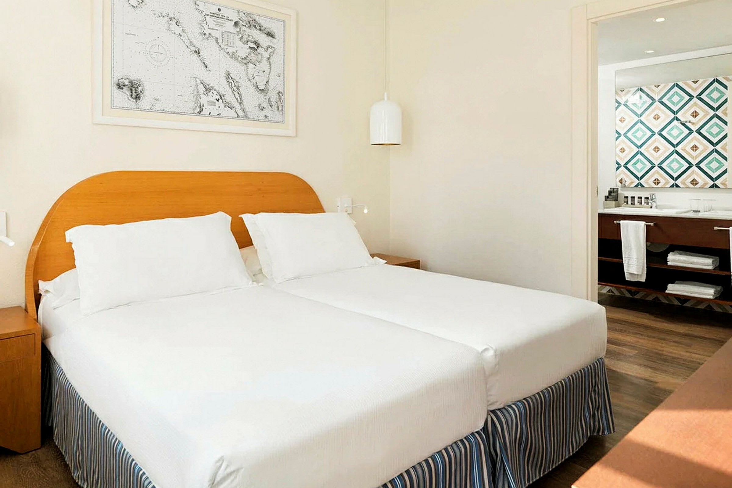En hotellrumsinteriör med två enkelsängar ihopställda, vita sängkläder och en ljus trästomme. En tavla hänger ovanför på väggen. I bakgrunden syns ett badrum med färgglada kakel. (AI-genererad text)