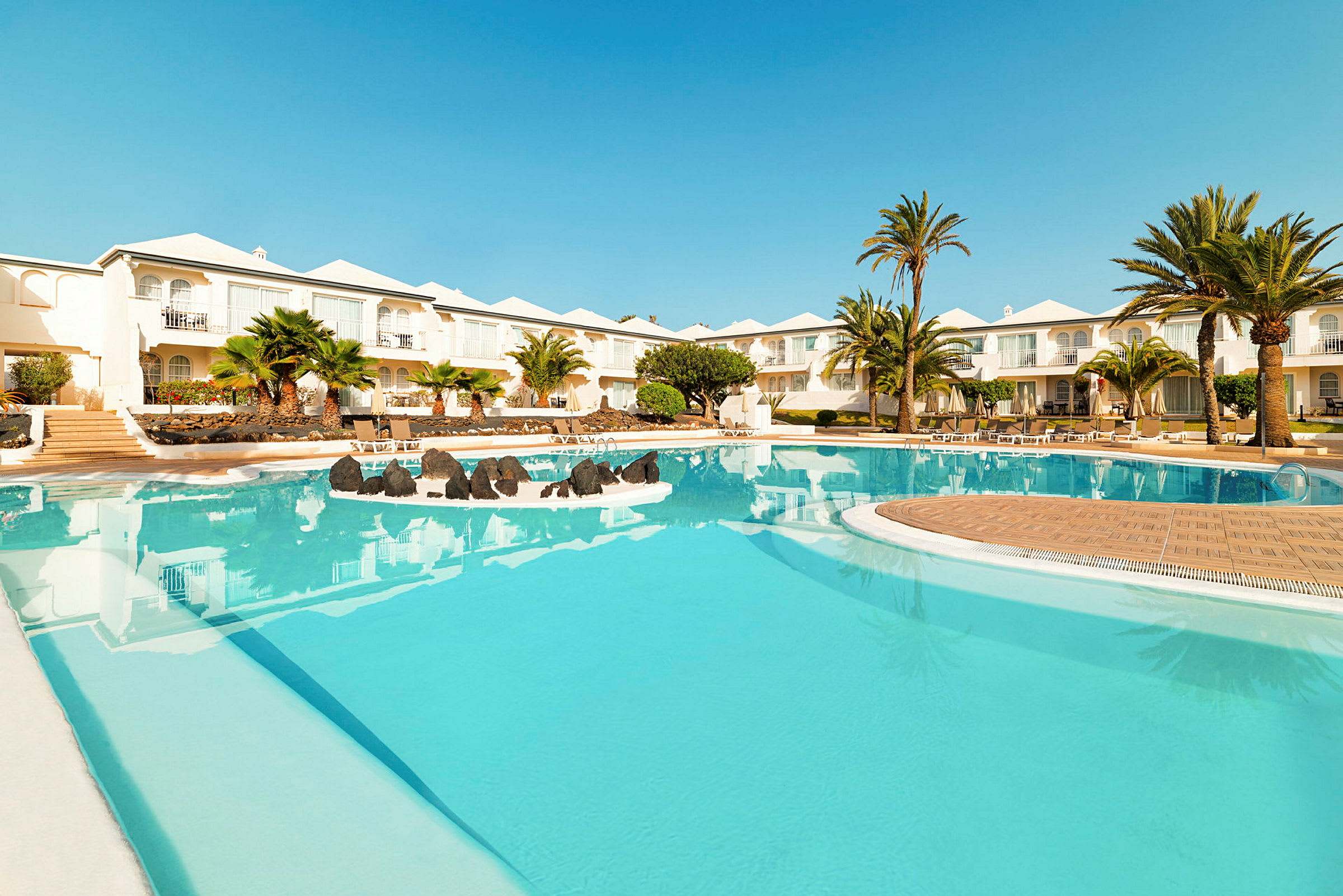 En bild på en hotellpool med klarblått vatten, omgärdad av palmträd och vita byggnader i bakgrunden. (AI-genererad text)