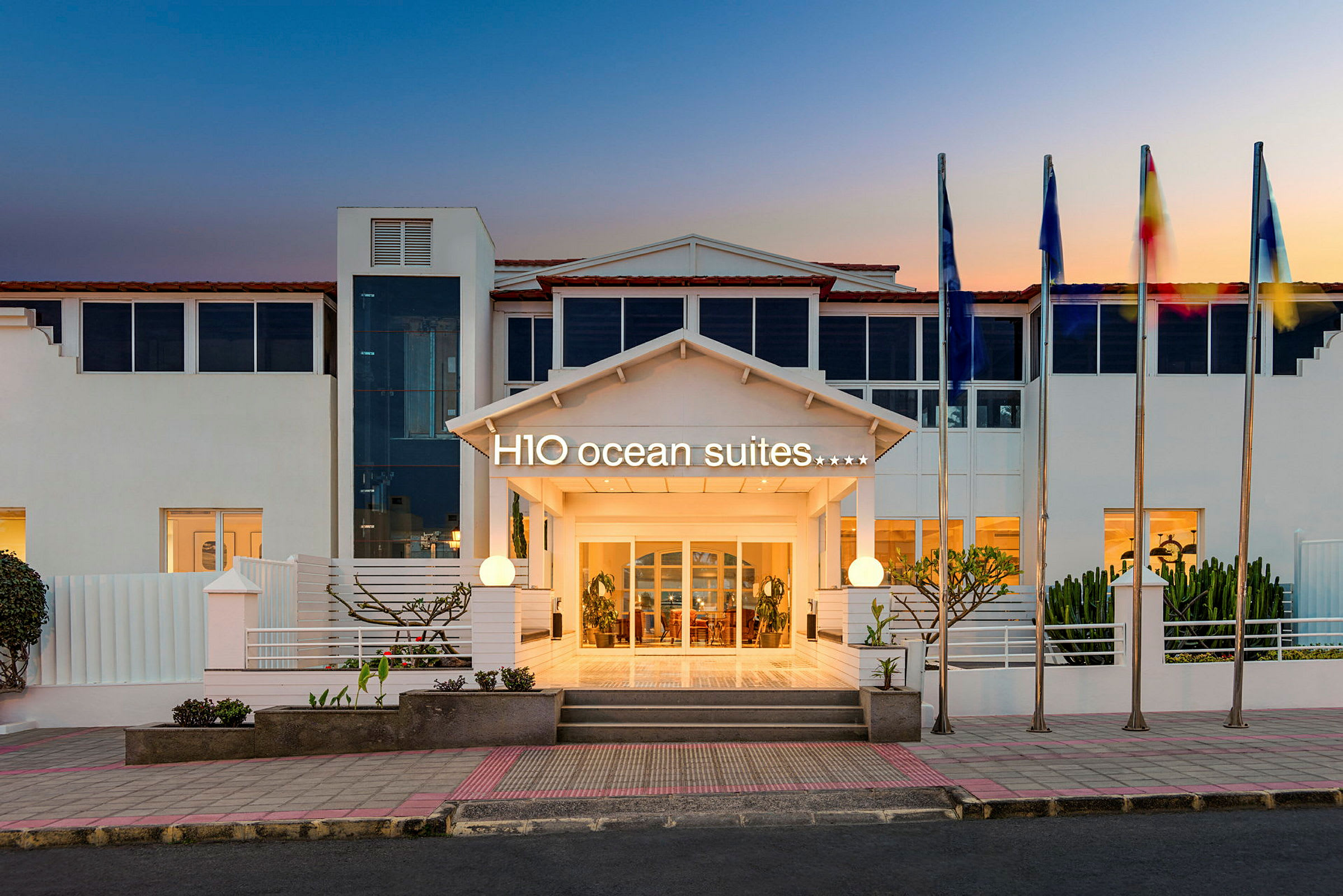 Hotellentré med skylten H10 Ocean Suites och flaggstänger vid solnedgång. (AI-genererad text)