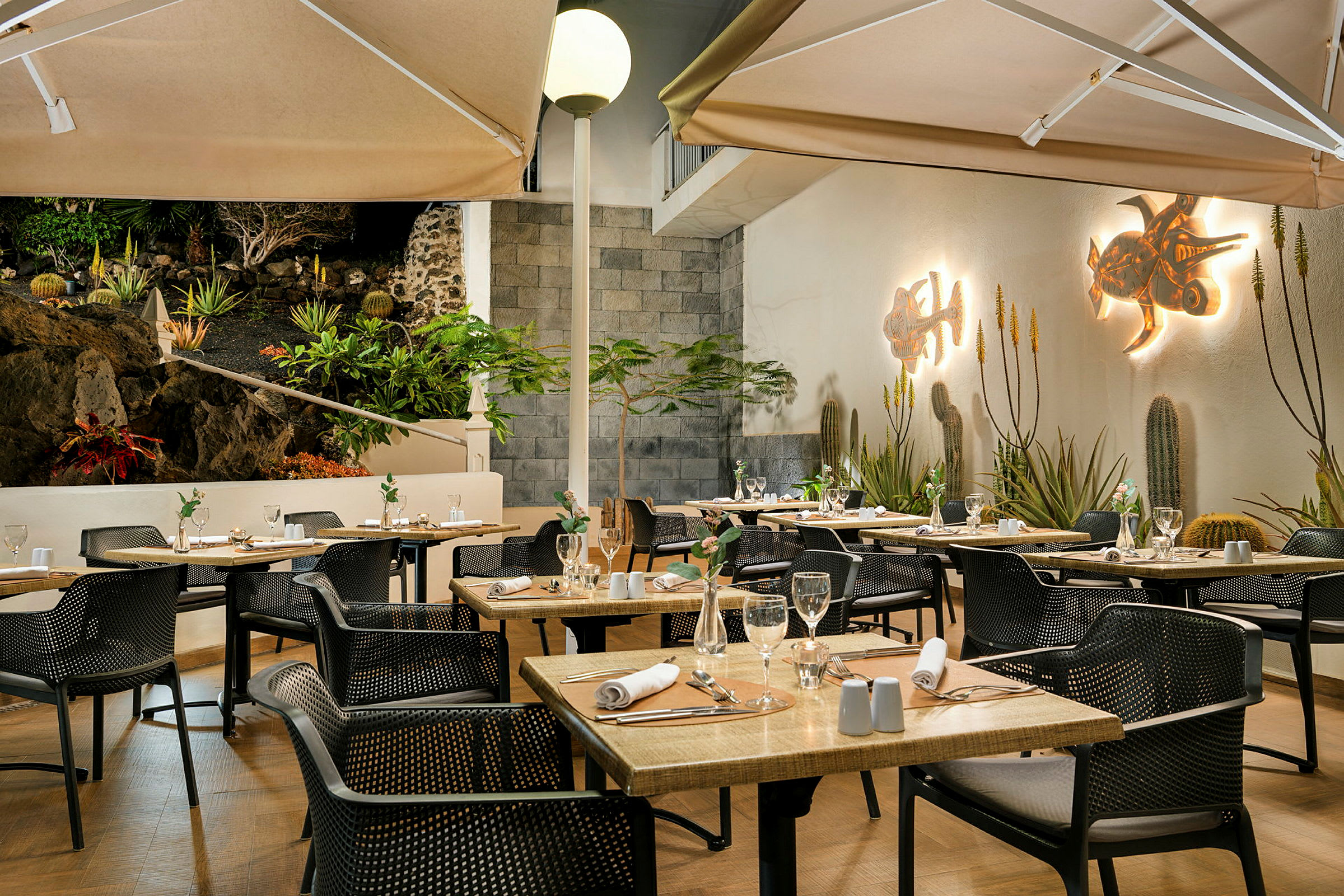 En väl upplyst restaurangterrass med dukade bord och stolar. Väggarna är dekorerade med konst och omgivningen har växter och en stenmur. (AI-genererad text)