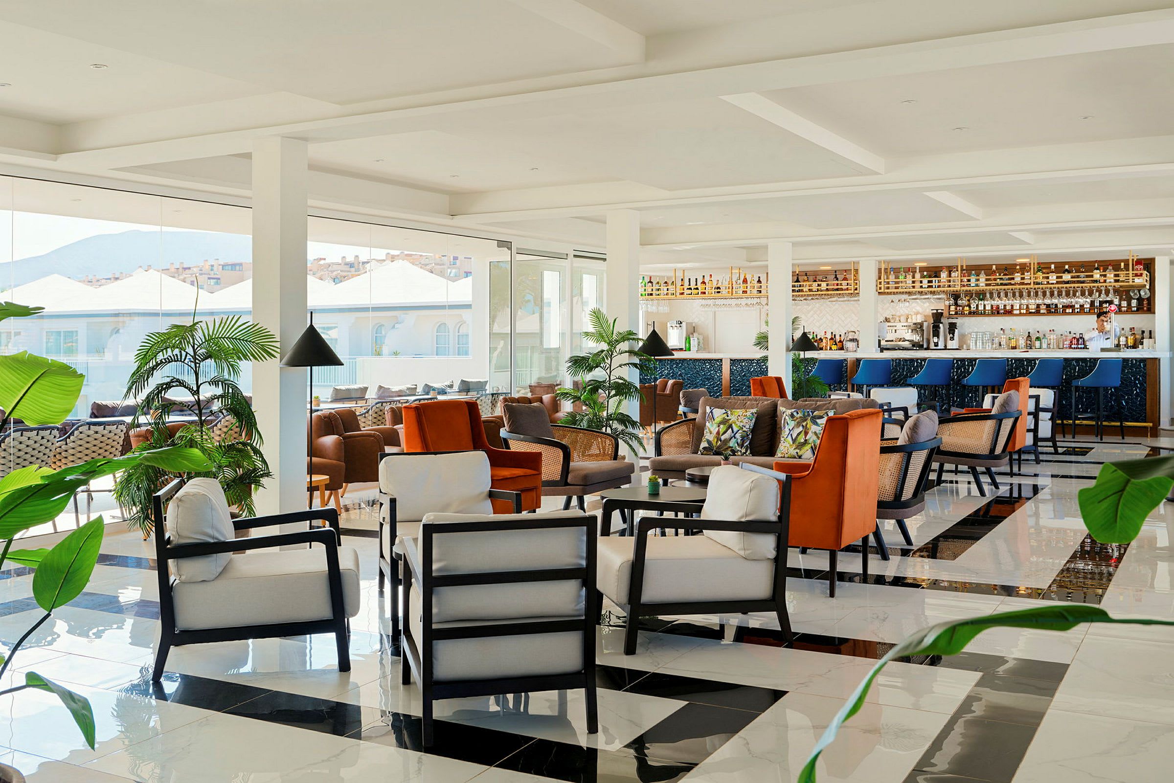 En modern lounge med bekväma sittmöbler, en bar och grönskande växter, samt stora fönster med utsikt mot byggnader och berg. (AI-genererad text)