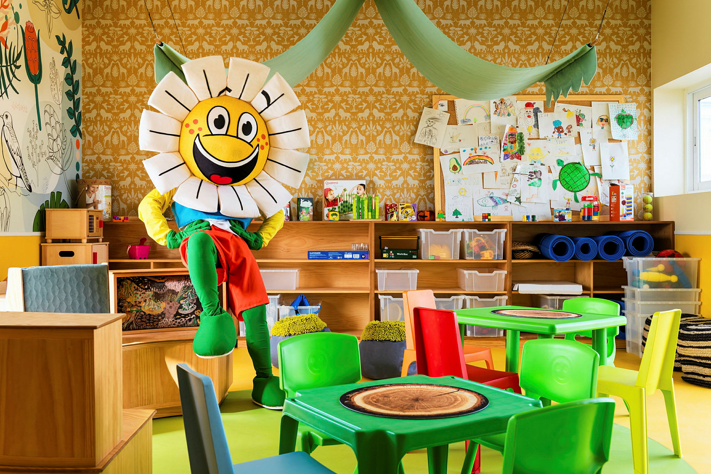Ett färgglatt rum med en maskot i form av en blomma som sitter vid möbler. Bakgrunden visar hyllor, förvaringslådor och teckningar. (AI-genererad text)