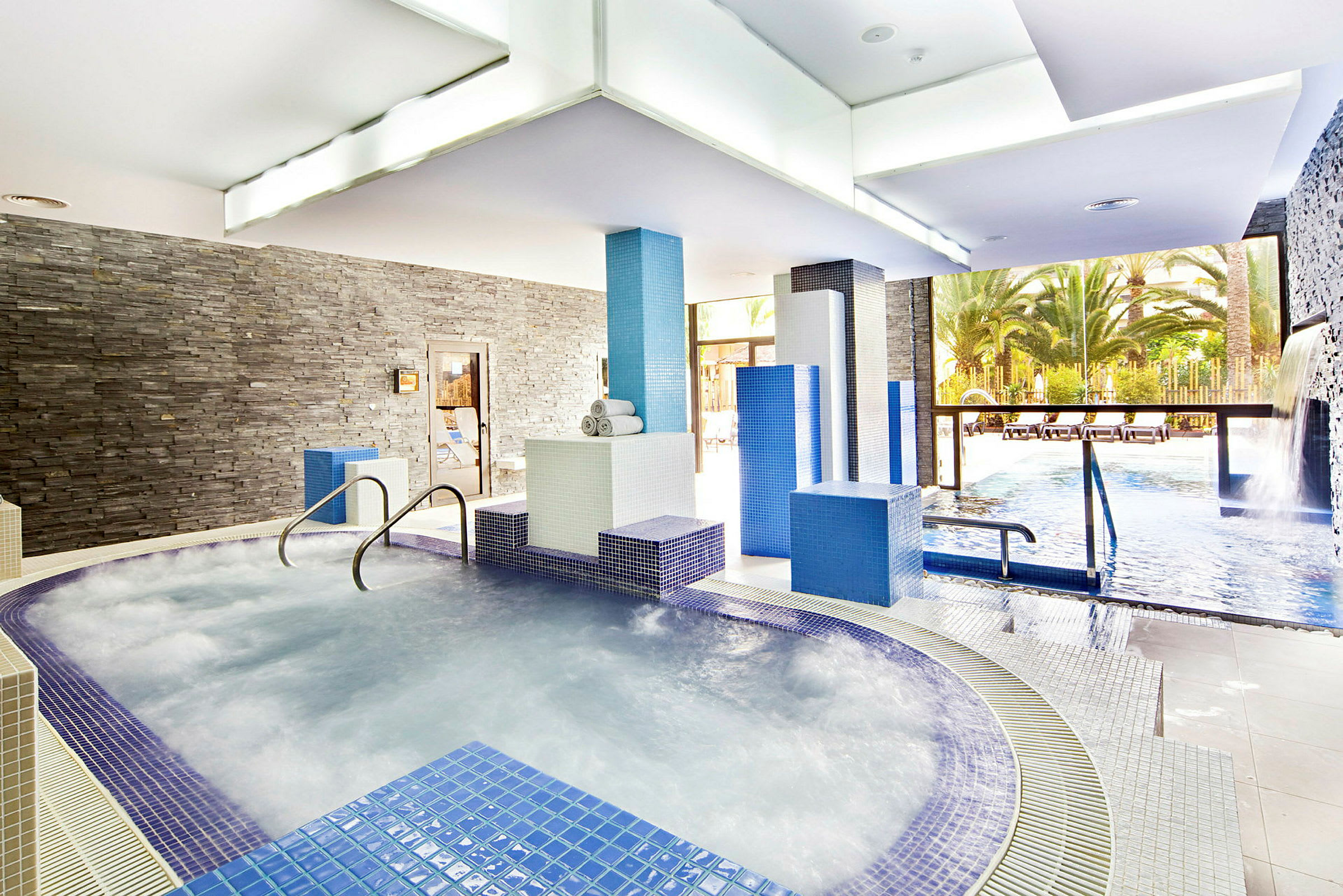 En interiör av ett spaområde med bubbelpool och pool omgiven av blå och vita kakel. Fönster öppnar upp mot en utomhuspool och palmer. (AI-genererad text)