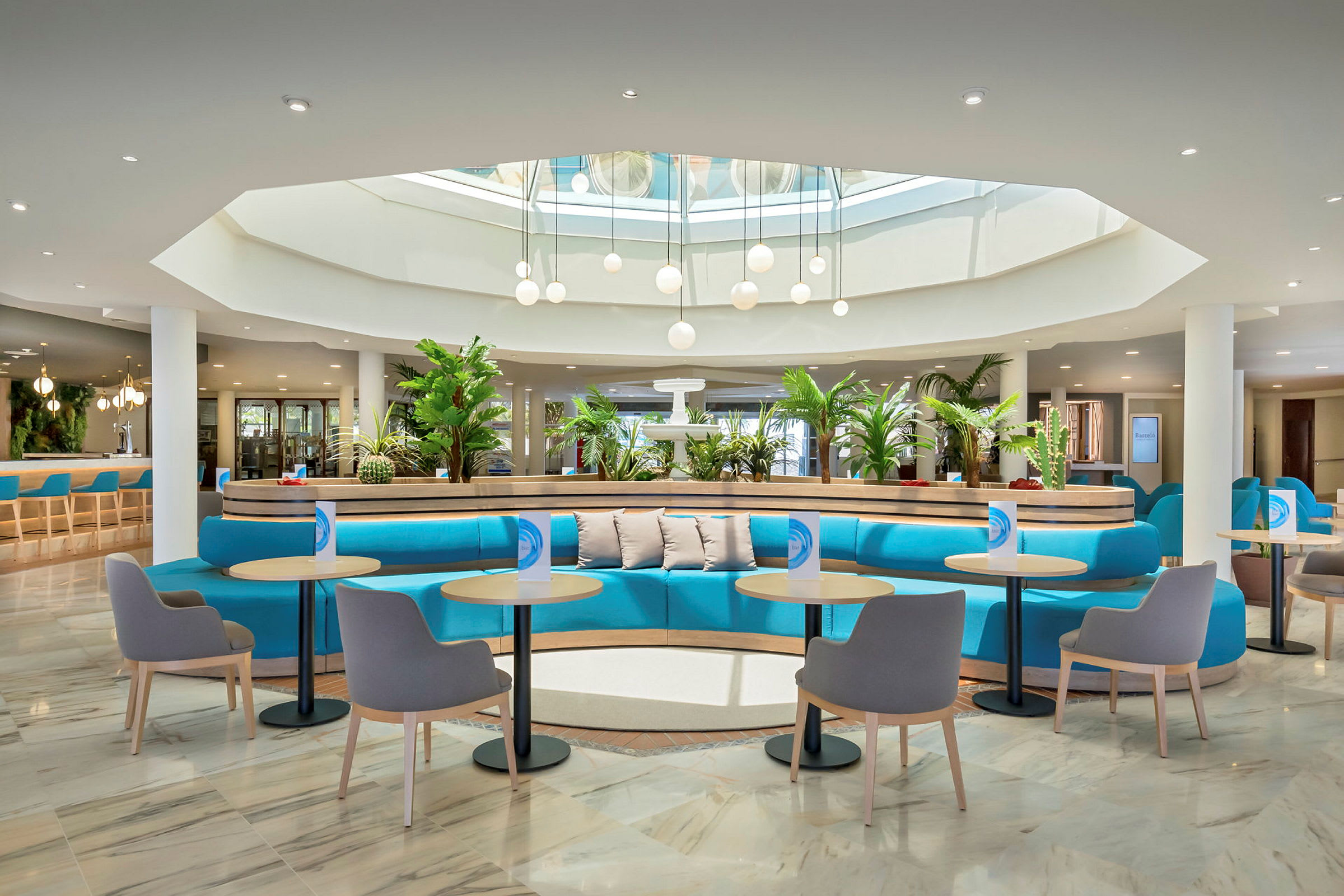 En modern hotellobby med blå sittsoffor, små bord och stolar. Växter pryder utrymmet, och takbelysning med glaskupor syns under ett takfönster. (AI-genererad text)