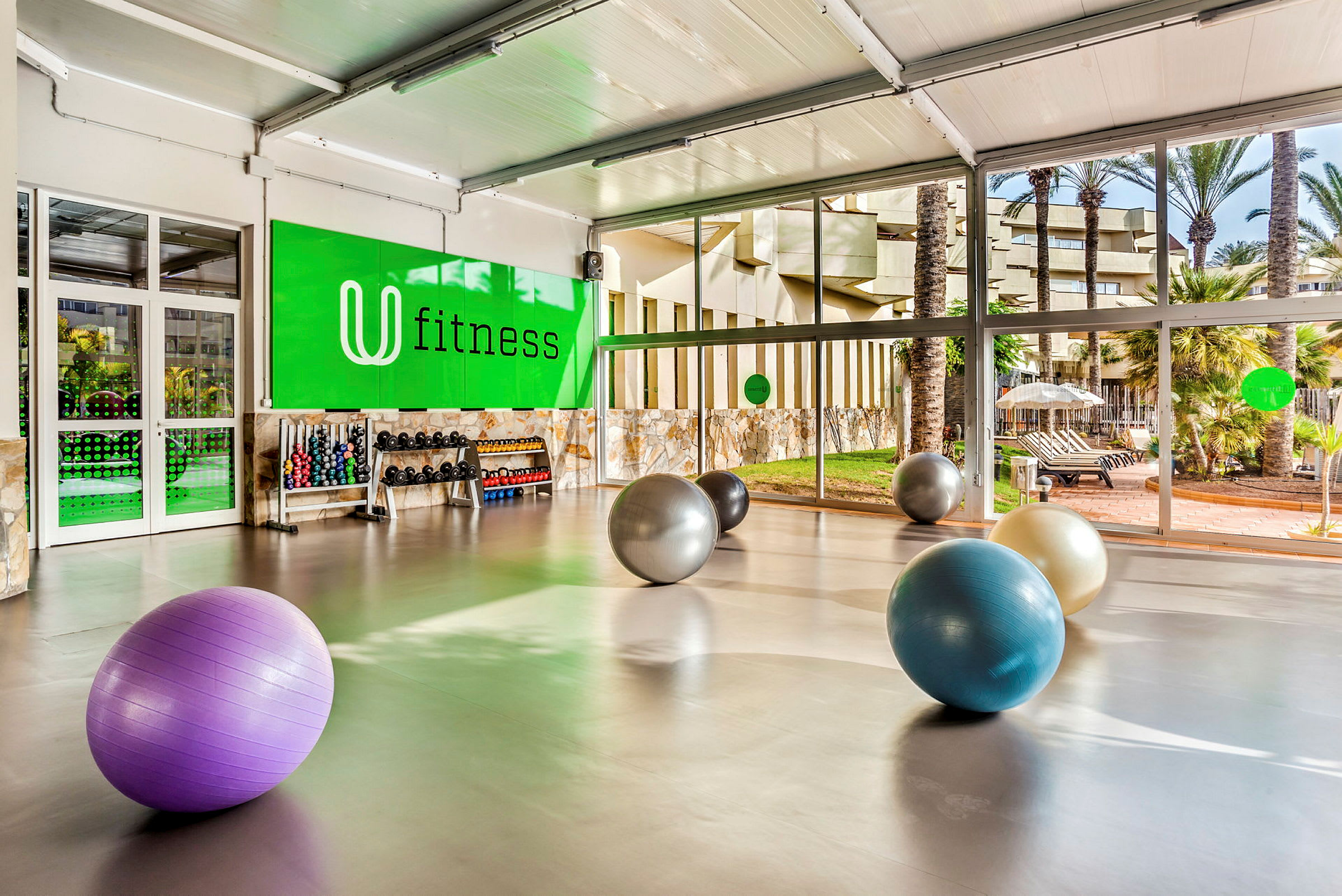 Ett gym med stora fönster och fitnessbollar på golvet. Dumbbellställ och träningsutrustning syns mot en grön vägg märkt "fitness". (AI-genererad text)