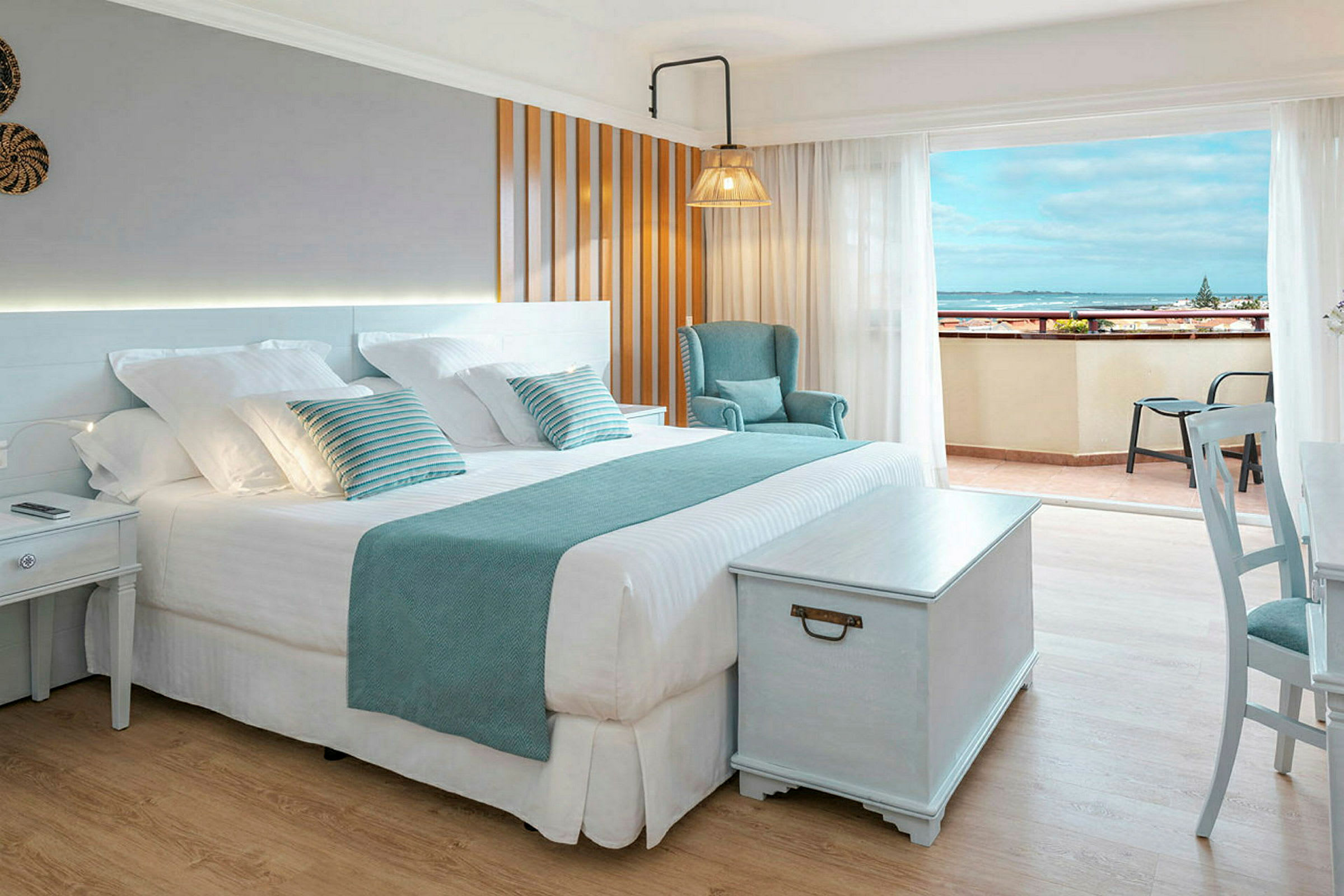 Ett ljust hotellrum med en stor dubbelsäng, stol och ett bord, samt en balkong med utsikt över havet. (AI-genererad text)