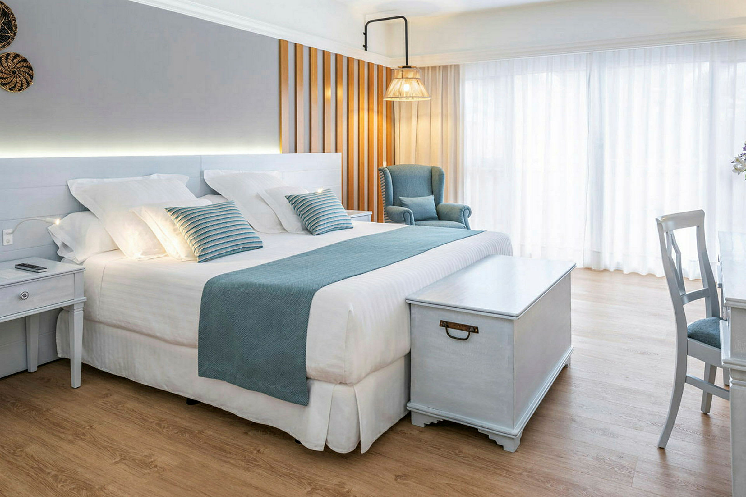 Ett ljust och elegant hotellrum med en stor säng, blå dekorationer, en fåtölj och ett träbord. (AI-genererad text)