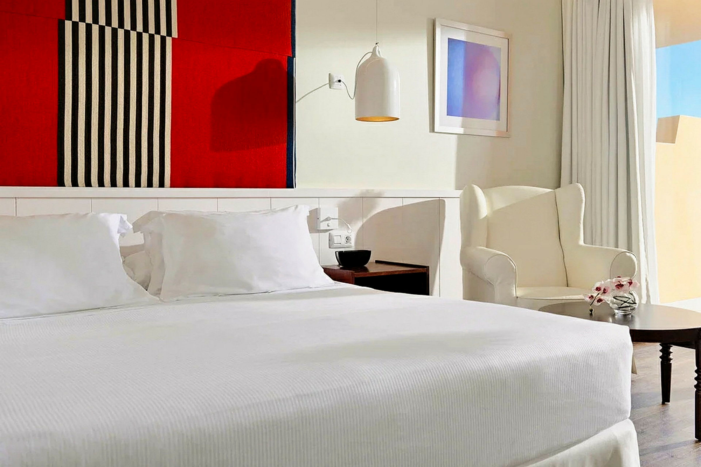 Ett stilrent hotellrum med en stor säng och vita sängkläder, en fåtölj bredvid ett litet bord, samt en färgglad textilväggdekoration ovanför sängen. (AI-genererad text)