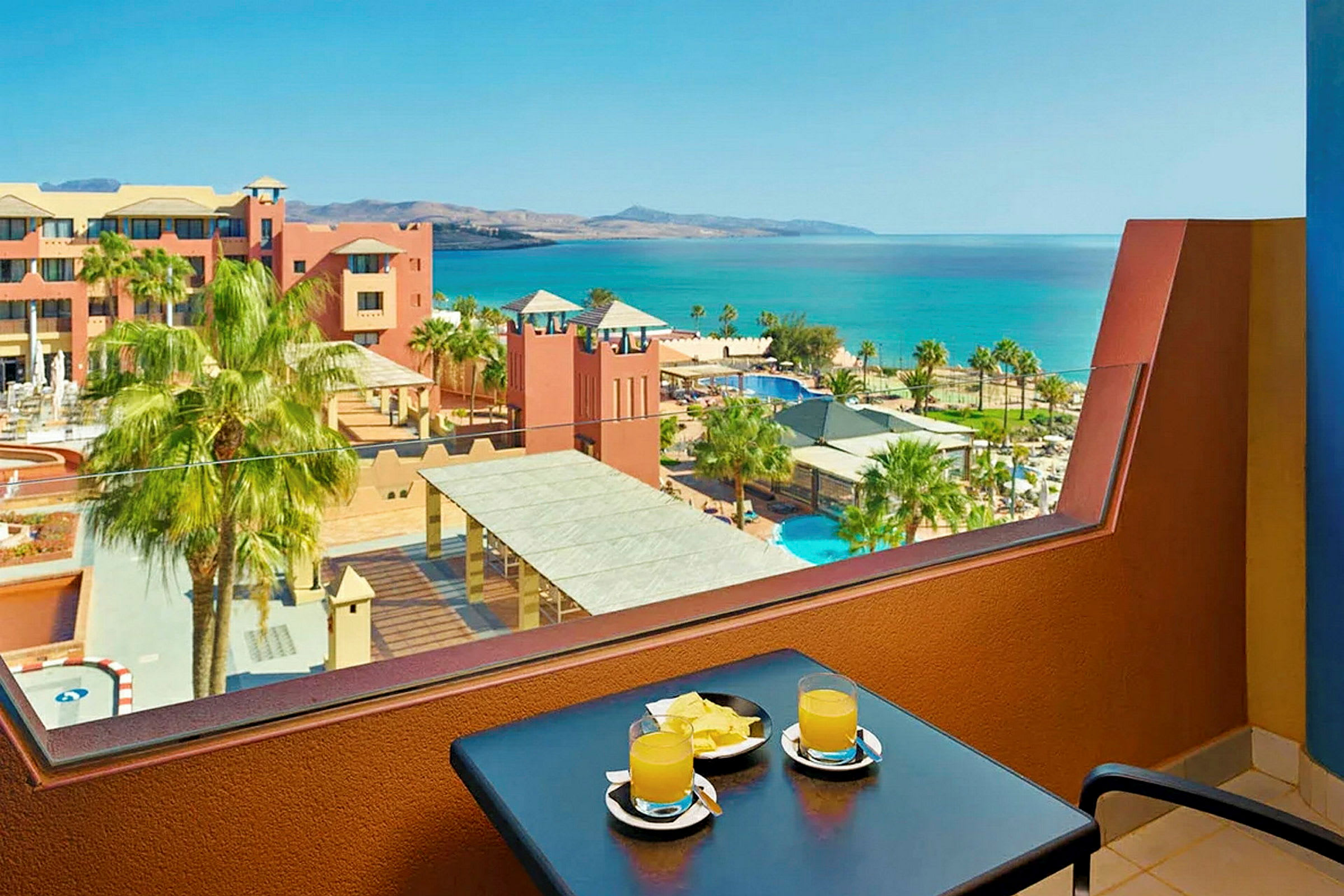 Utsikt från en balkong med ett bord där två glas med juice och några bitar av frukt är placerade. I bakgrunden syns en resort med pooler, palmer och havet. (AI-genererad text)