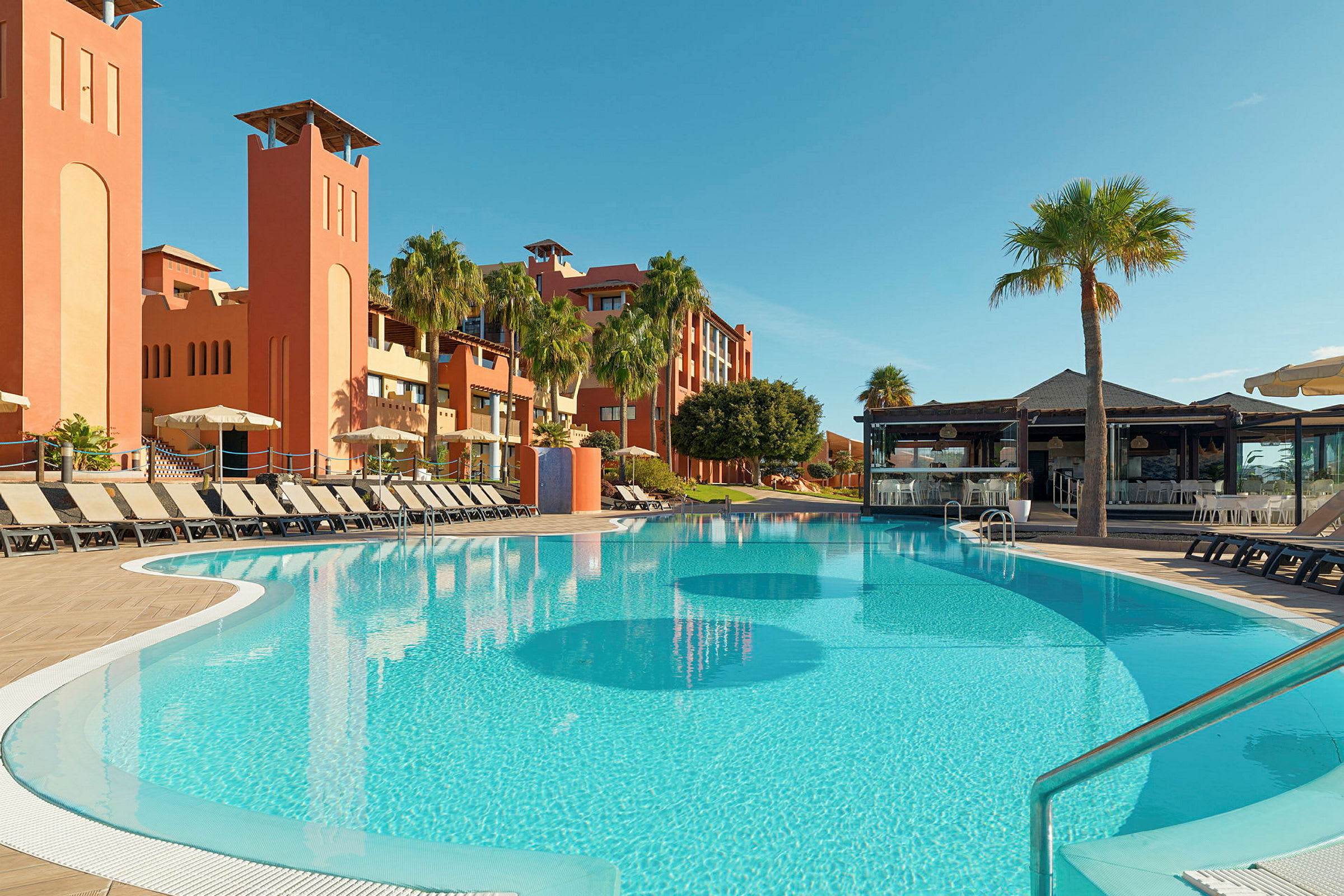 Utomhuspool med solstolar i ett resortområde, omgiven av palmer och orange byggnader. (AI-genererad text)
