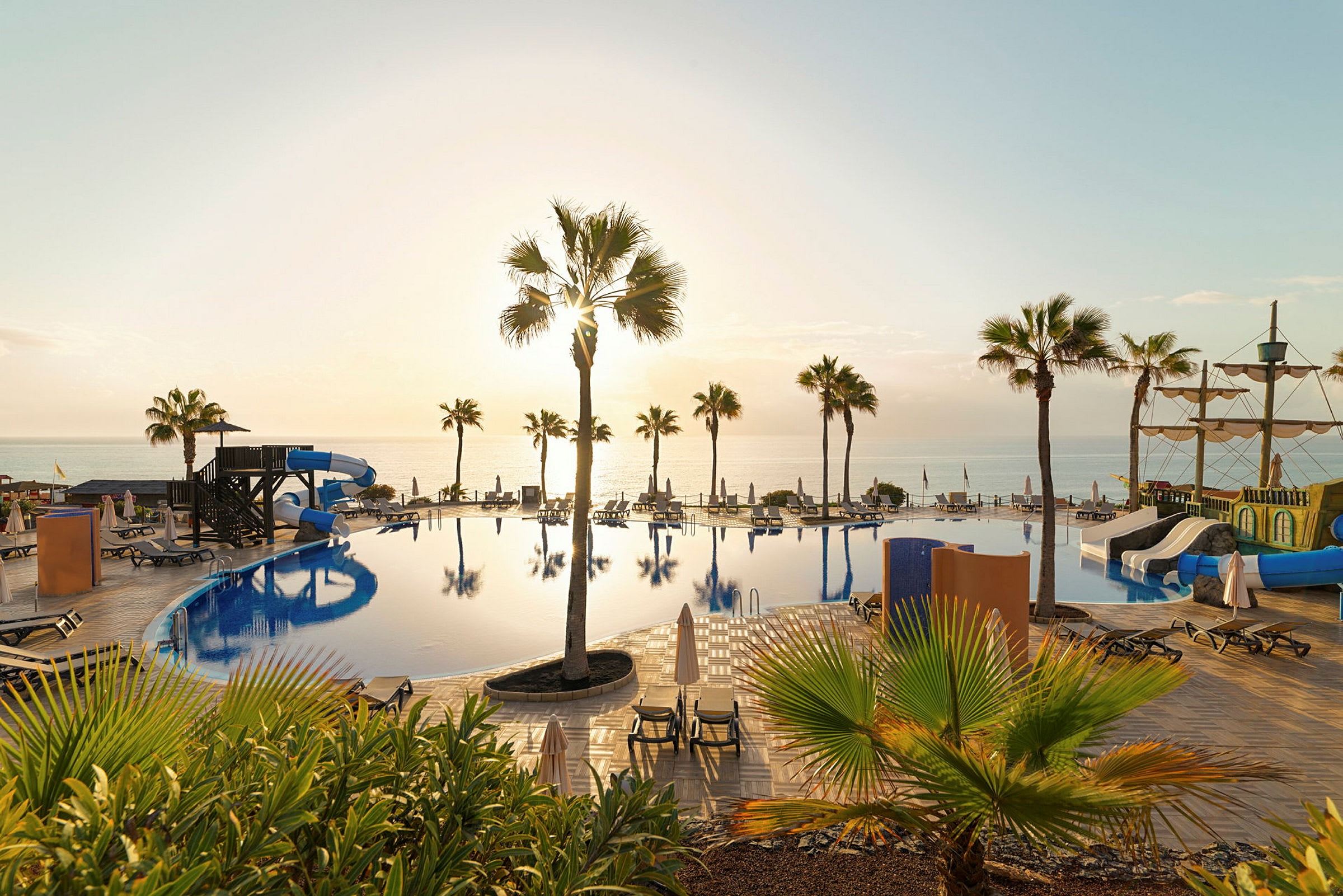 Bild på en pool med vattenrutschbanor, solstolar och palmer, samt havet i bakgrunden vid solnedgång. (AI-genererad text)