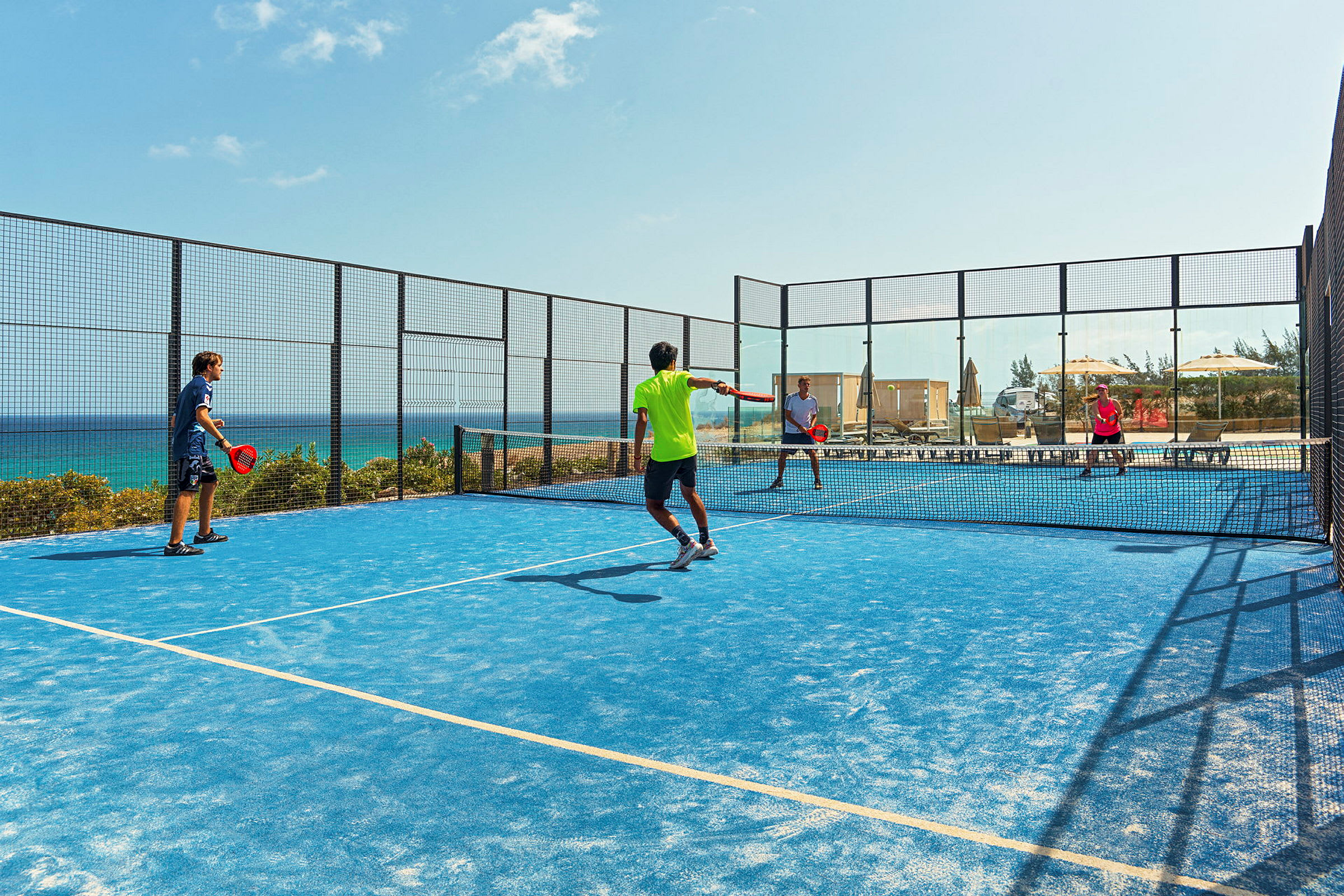 Fyra personer spelar padel på en utomhusbana med blått underlag. Havet syns i bakgrunden. (AI-genererad text)