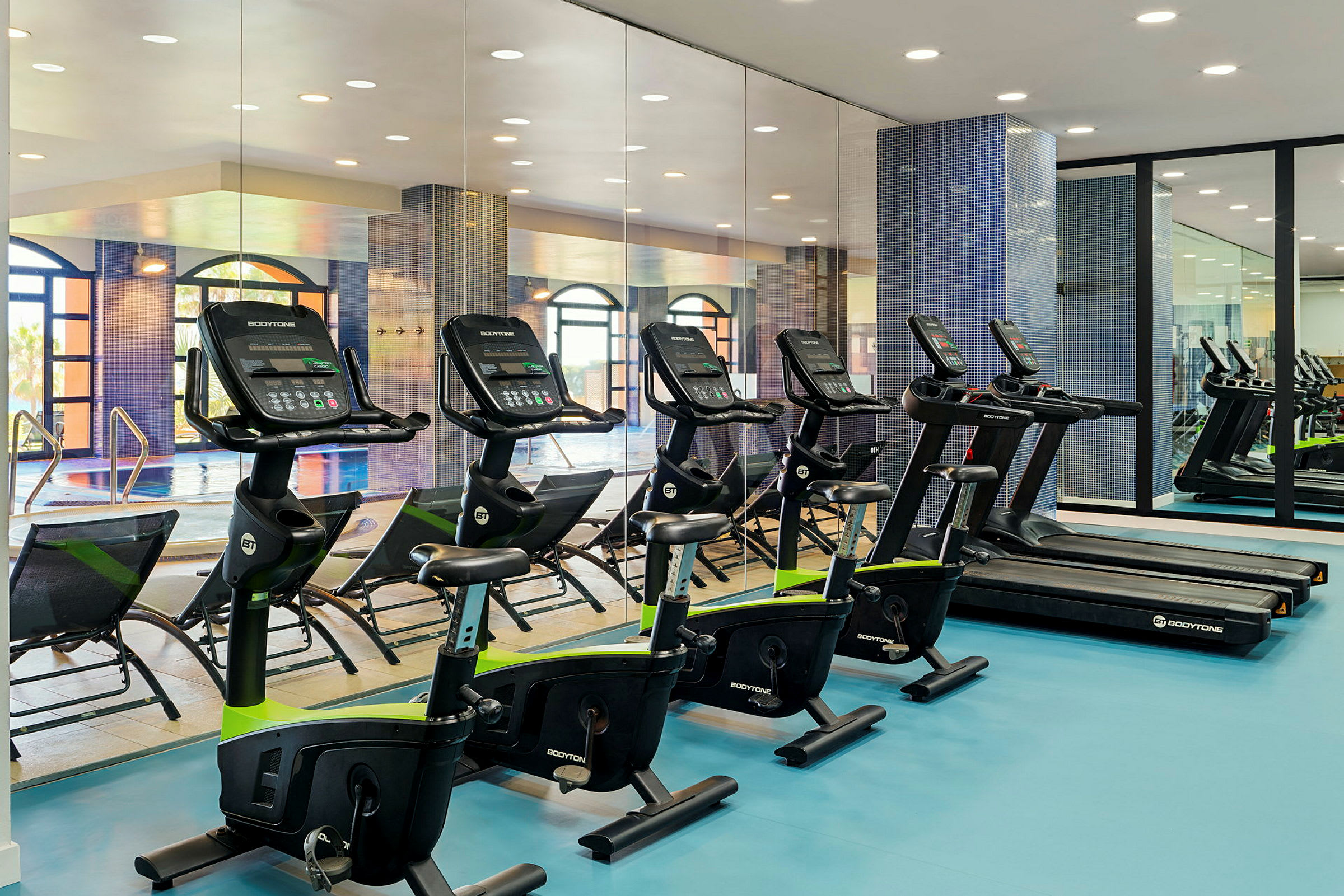 Bild av ett gym med träningscyklar och löpband placerade på rad. I bakgrunden syns en spegelvägg och en swimmingpool. (AI-genererad text)