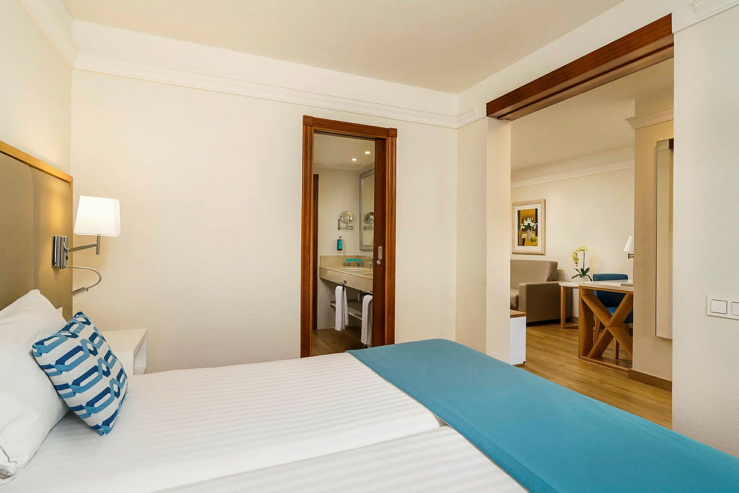 Ett hotellrum med ett sängområde i förgrunden, badrum med spegel synlig genom en dörröppning i mitten, och ett vardagsrum med sittgrupp i bakgrunden. (AI-genererad text)