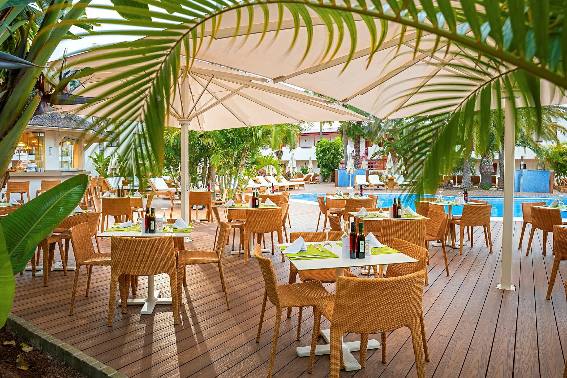 Utomhusrestaurang med trätrall, bord med dukar och stolar, omgivet av grönska och en pool. (AI-genererad text)