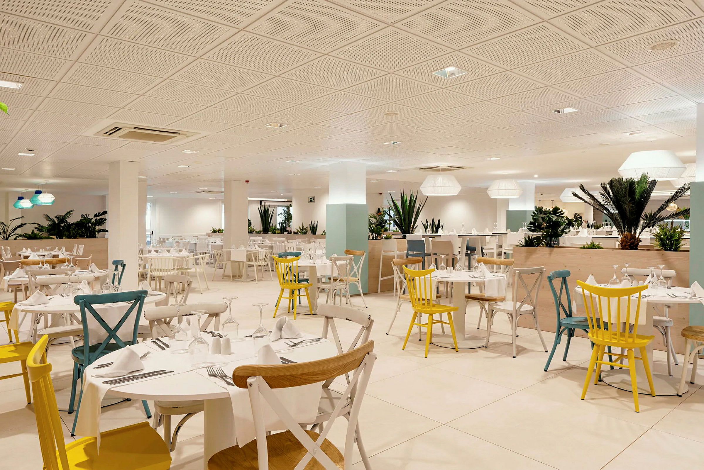 Interiören av en ljus restaurang med många bord och stolar i olika färger, dekorerad med gröna växter. (AI-genererad text)