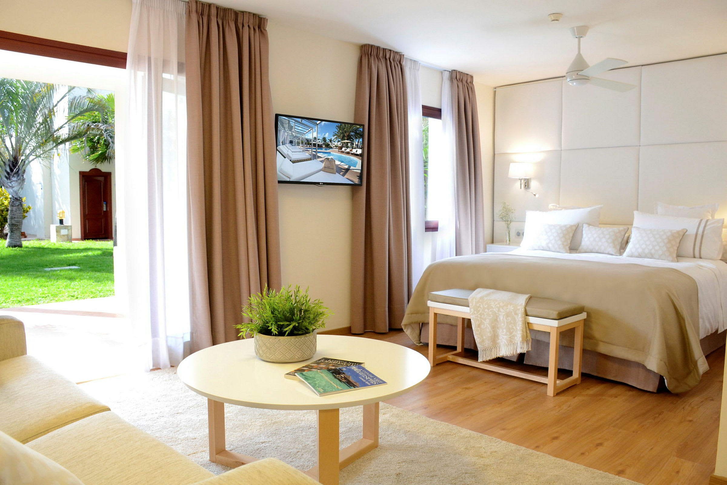 Ett ljust hotellrum med en stor säng, väggmonterad TV, och en sittgrupp framför en rund bord. Stora gardiner med utsikt mot en trädgård. (AI-genererad text)