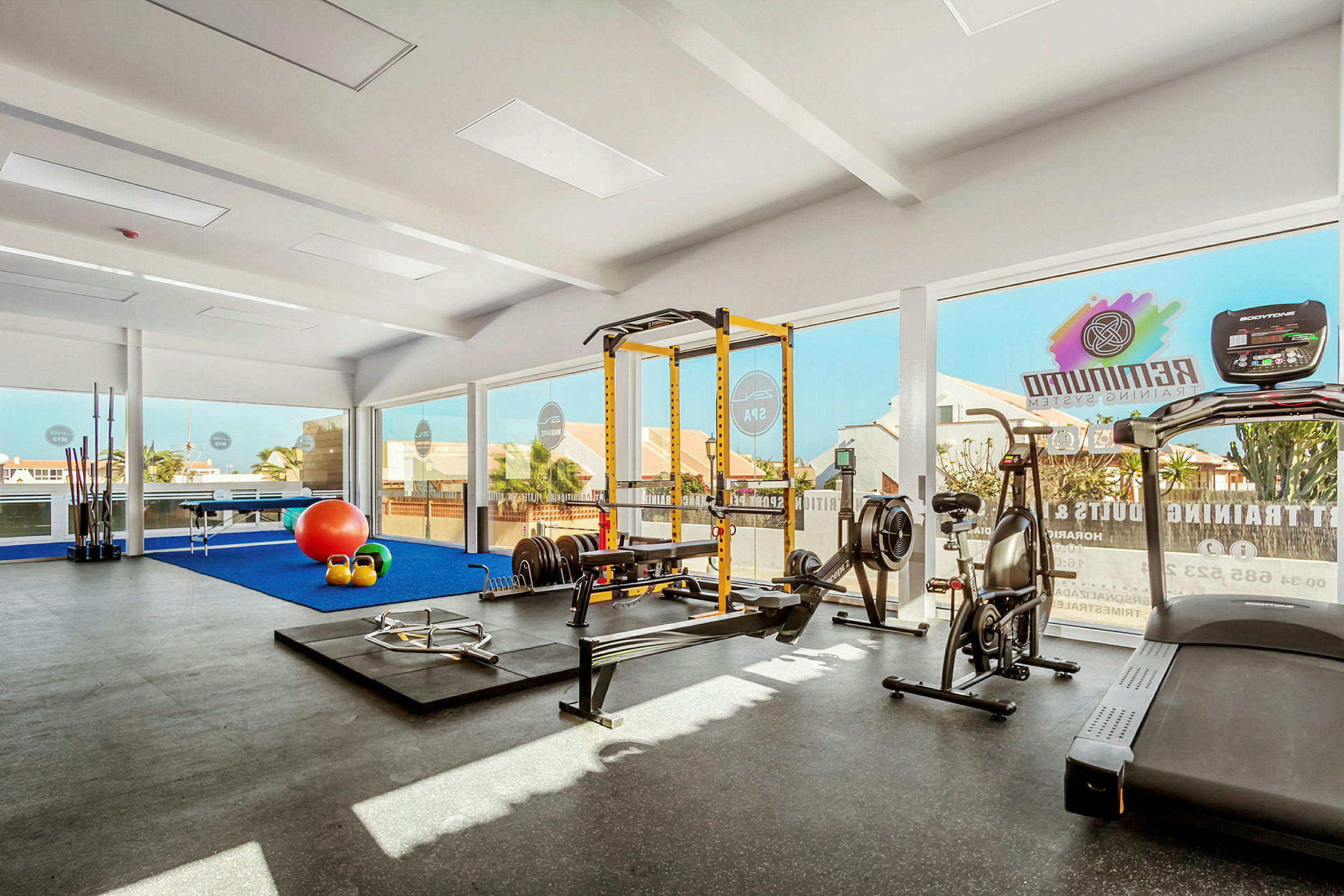 Ett gym med träningsutrustning som inkluderar löpband, roddmaskin, vikter och en träningsställning. Stora fönster släpper in dagsljus. (AI-genererad text)