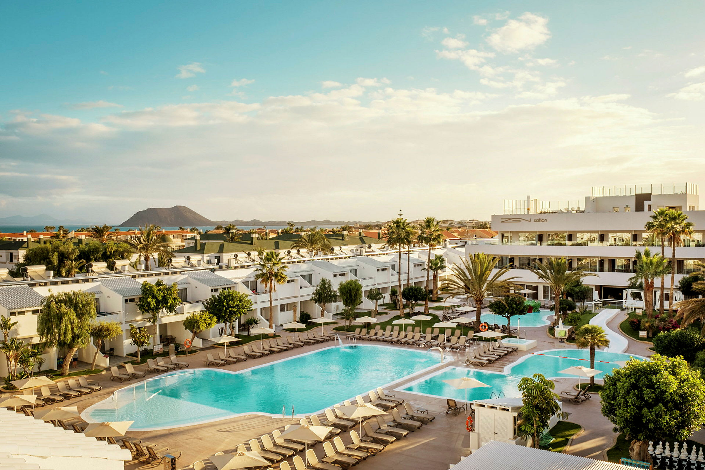 Bild av ett hotellområde med pooler, solstolar och omgivande byggnader, samt en vulkan i bakgrunden. (AI-genererad text)