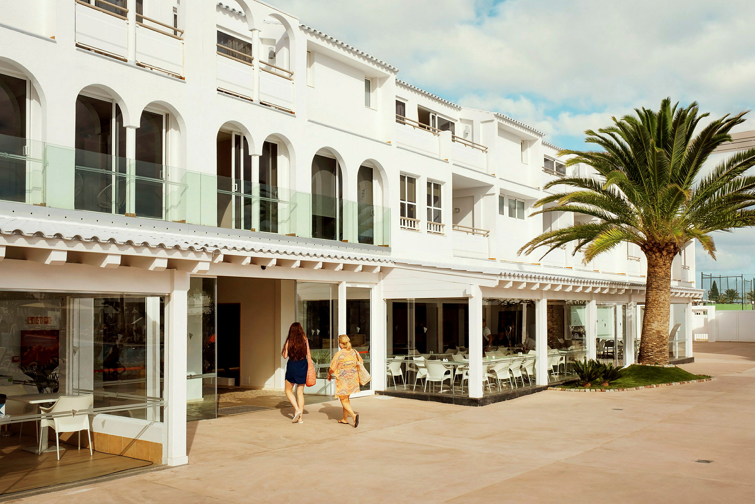 Ett vitt hotellkomplex med stora fönster och en restaurang med sittplatser utomhus. En palm står vid byggnaden och två personer går mot ingången. (AI-genererad text)