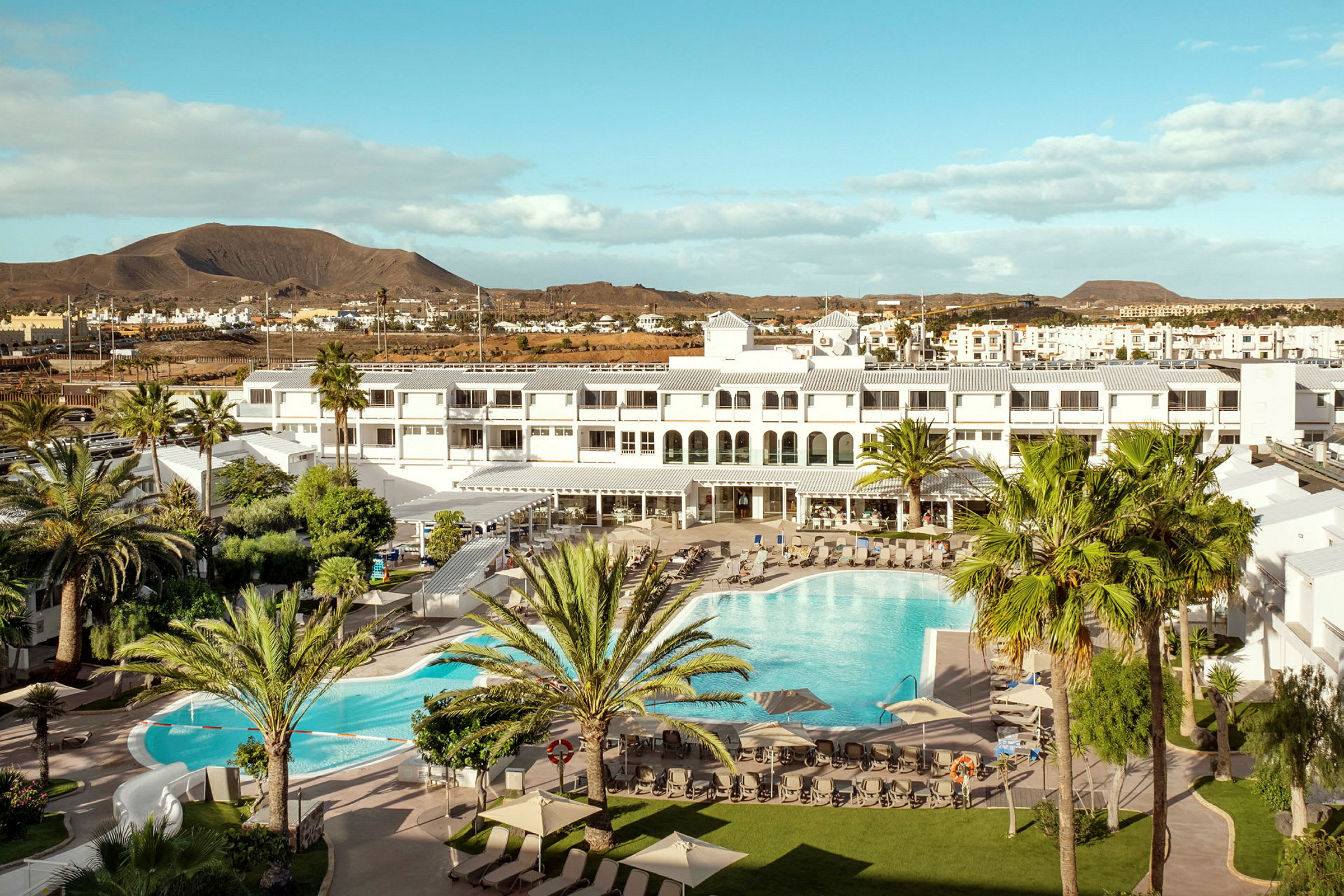 Ett hotellområde med en stor pool, omgiven av palmer och solstolar. I bakgrunden syns låga byggnader och ett berg. (AI-genererad text)