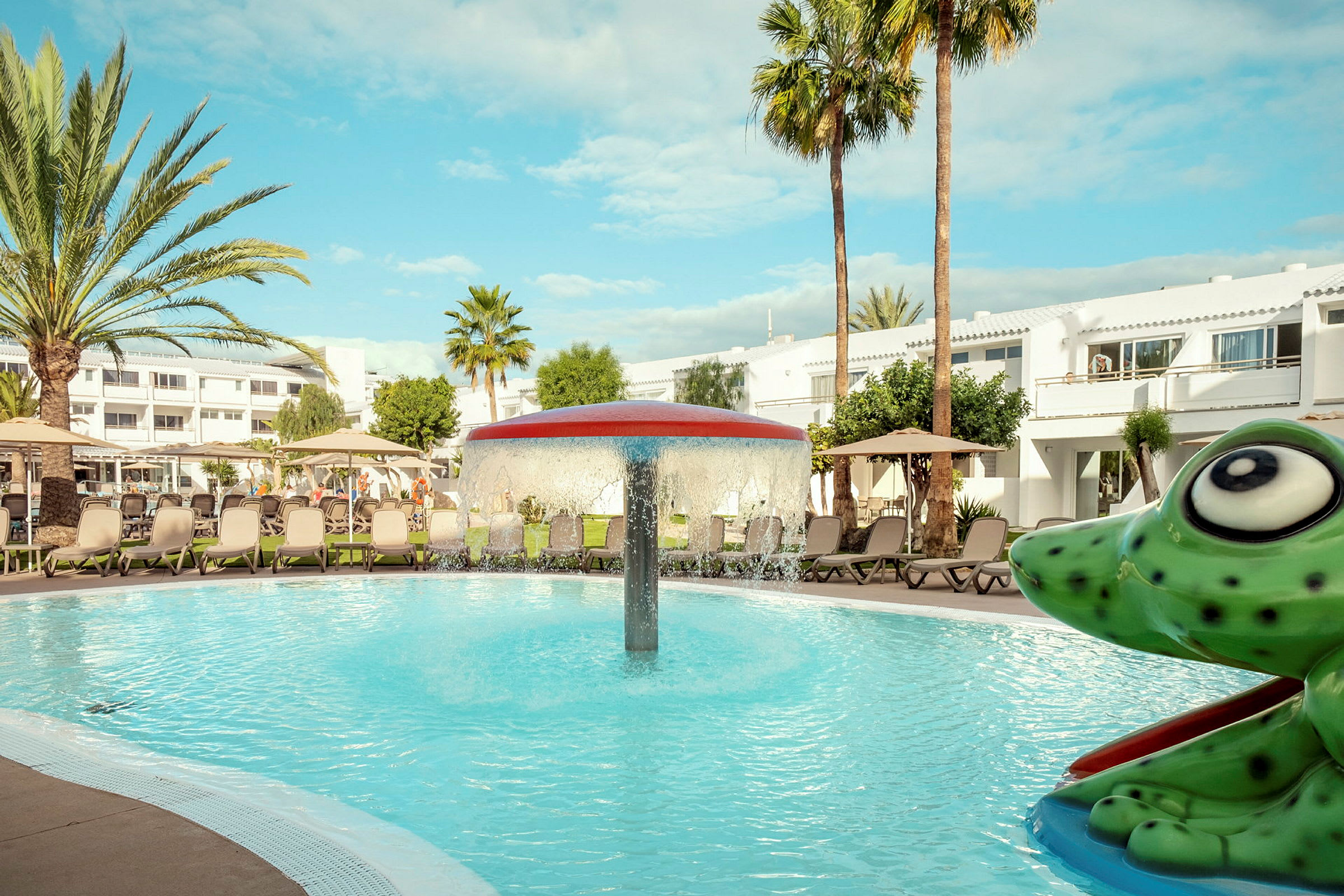 En pool med en svampformad fontän i mitten, omgiven av solstolar och parasoller. I förgrunden syns en figur formad som en grön groda. (AI-genererad text)