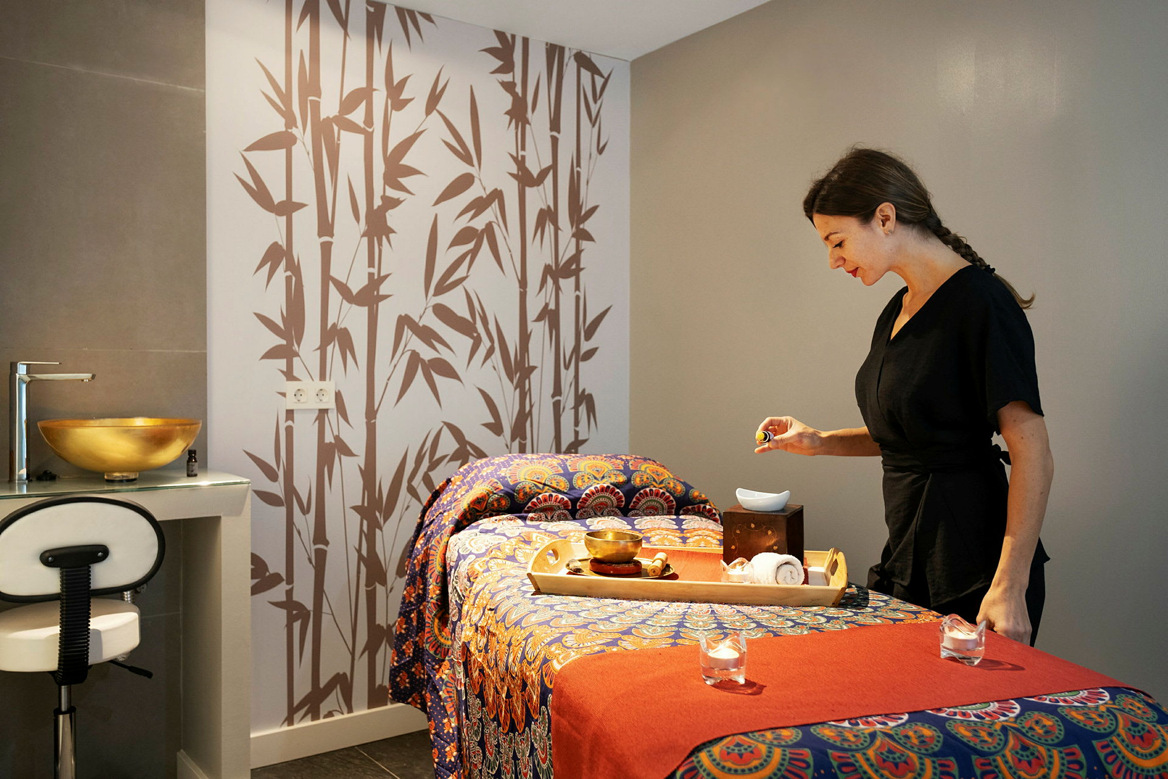 Ett spa- eller massagesrum med en dekorerad massagebänk, en bricka med tillbehör och en person vid sidan av. (AI-genererad text)