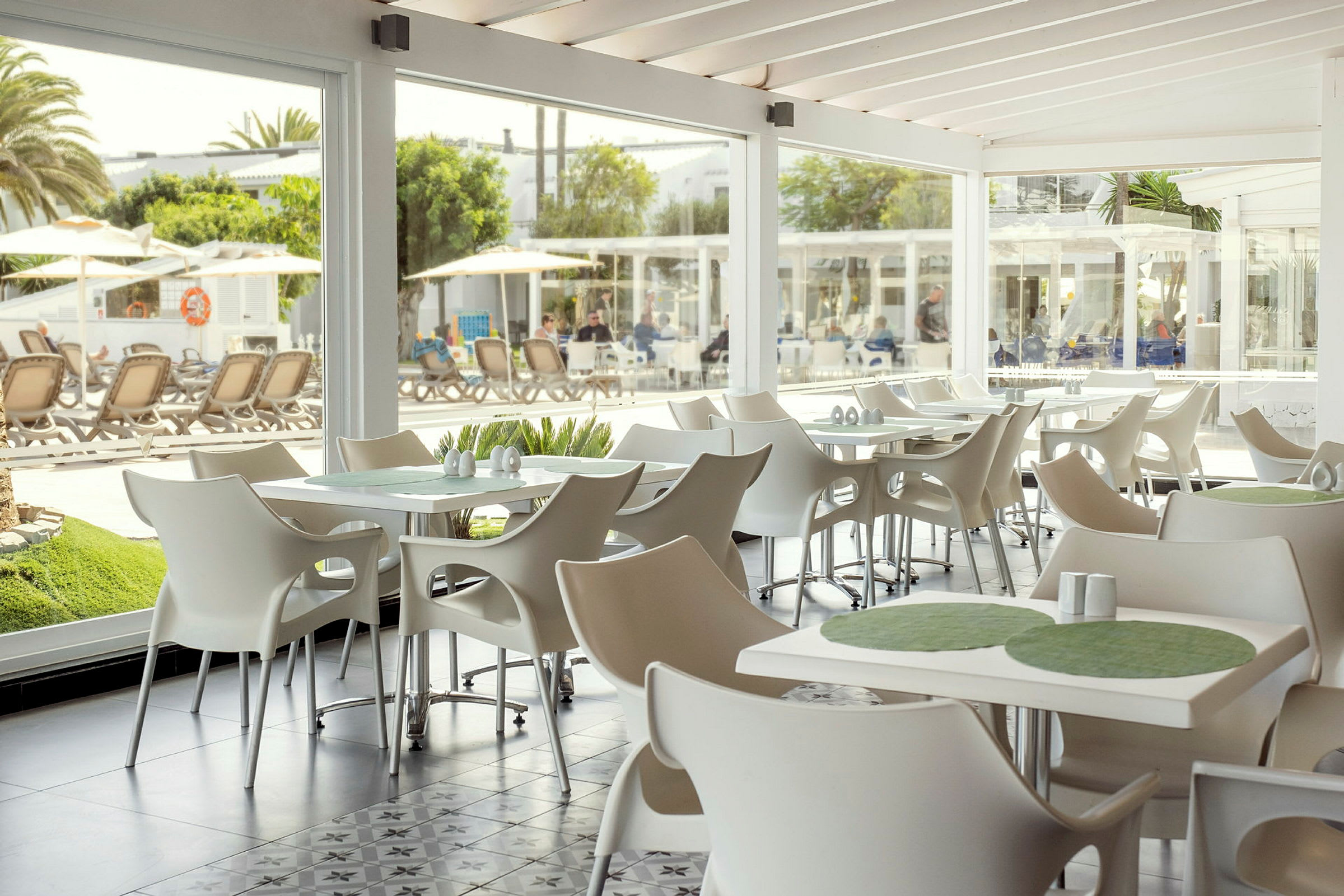 Ett ljust och modern inrett café med vita stolar och bord, med utsikt mot utomhuspoolområde genom stora glasfönster. (AI-genererad text)
