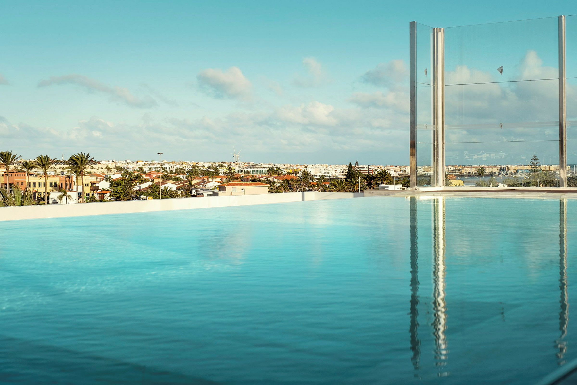 En infinitypool med utsikt över stadshus, palmer och en vindmölla under en blå himmel. (AI-genererad text)