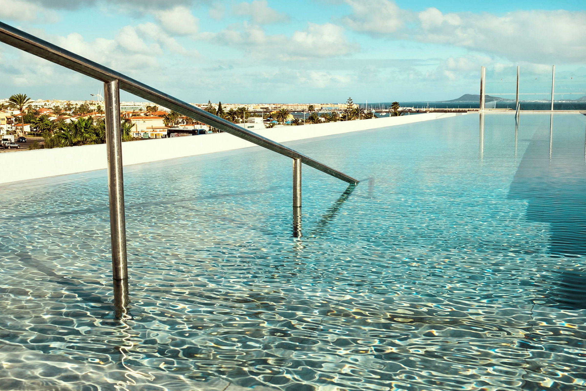 En pool med klart vatten och ett räcke som leder ner i vattnet. I bakgrunden syns en stad och blå himmel med några moln. (AI-genererad text)