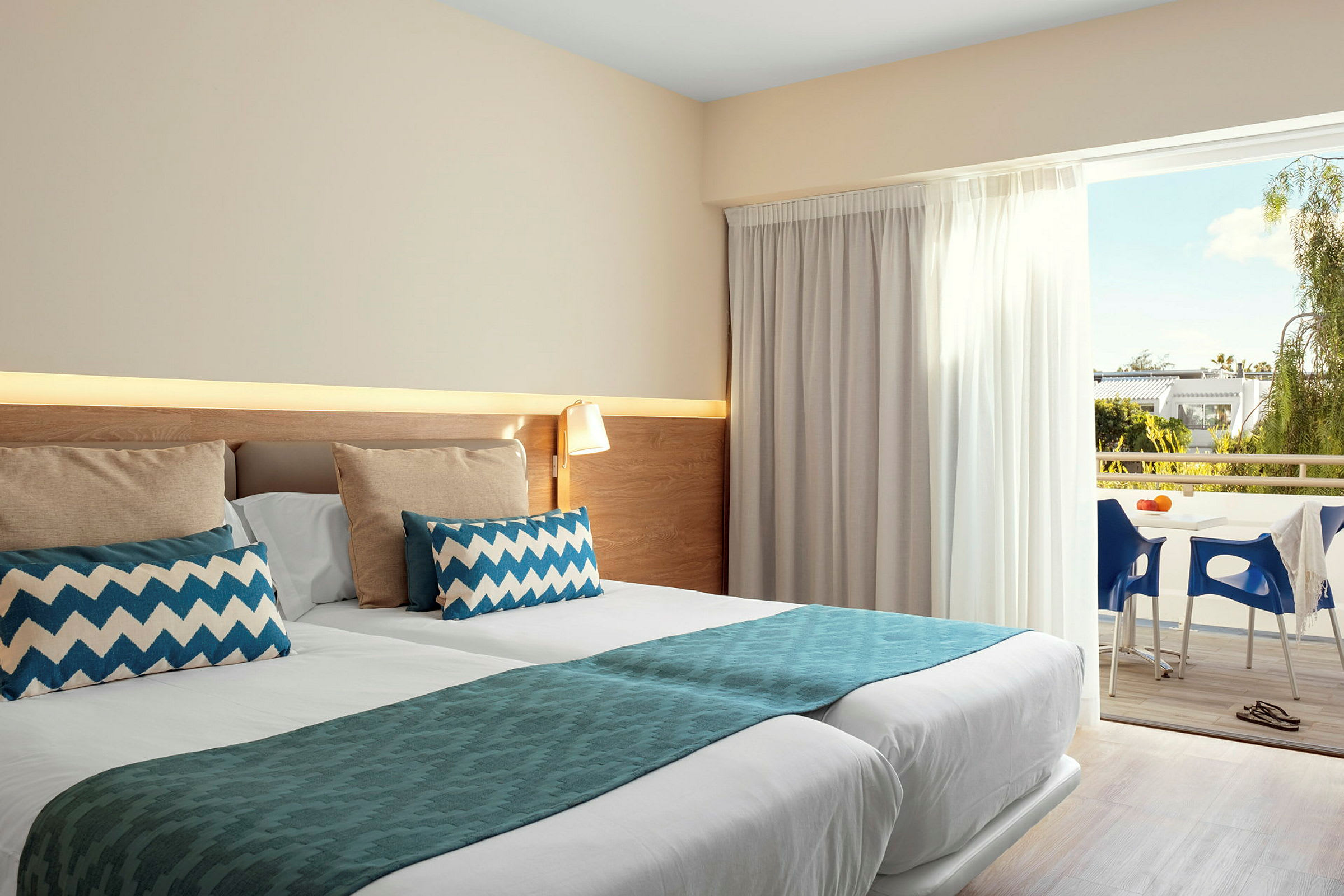Ett hotellrum med dubbelsäng som är snyggt bäddad med blå detaljer och kuddar. Rummet har även tillgång till en balkong med utsikt över grönska och möbler. (AI-genererad text)
