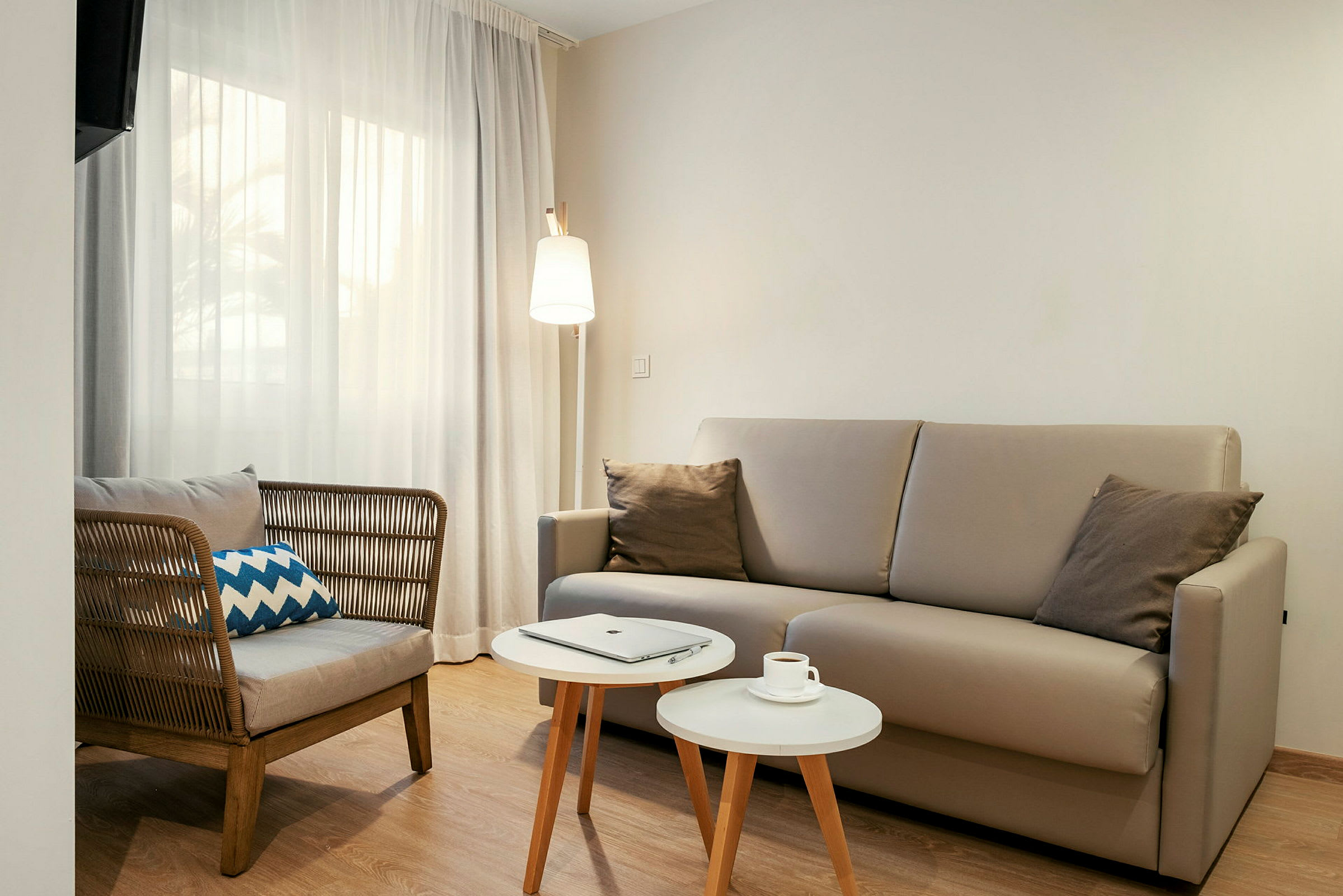 Ett vardagsrum med en beige soffa, en stol med rygg av flätat material, två vita små bord och en golvlampa. Ett fönster med ljusa gardiner syns i bakgrunden. (AI-genererad text)