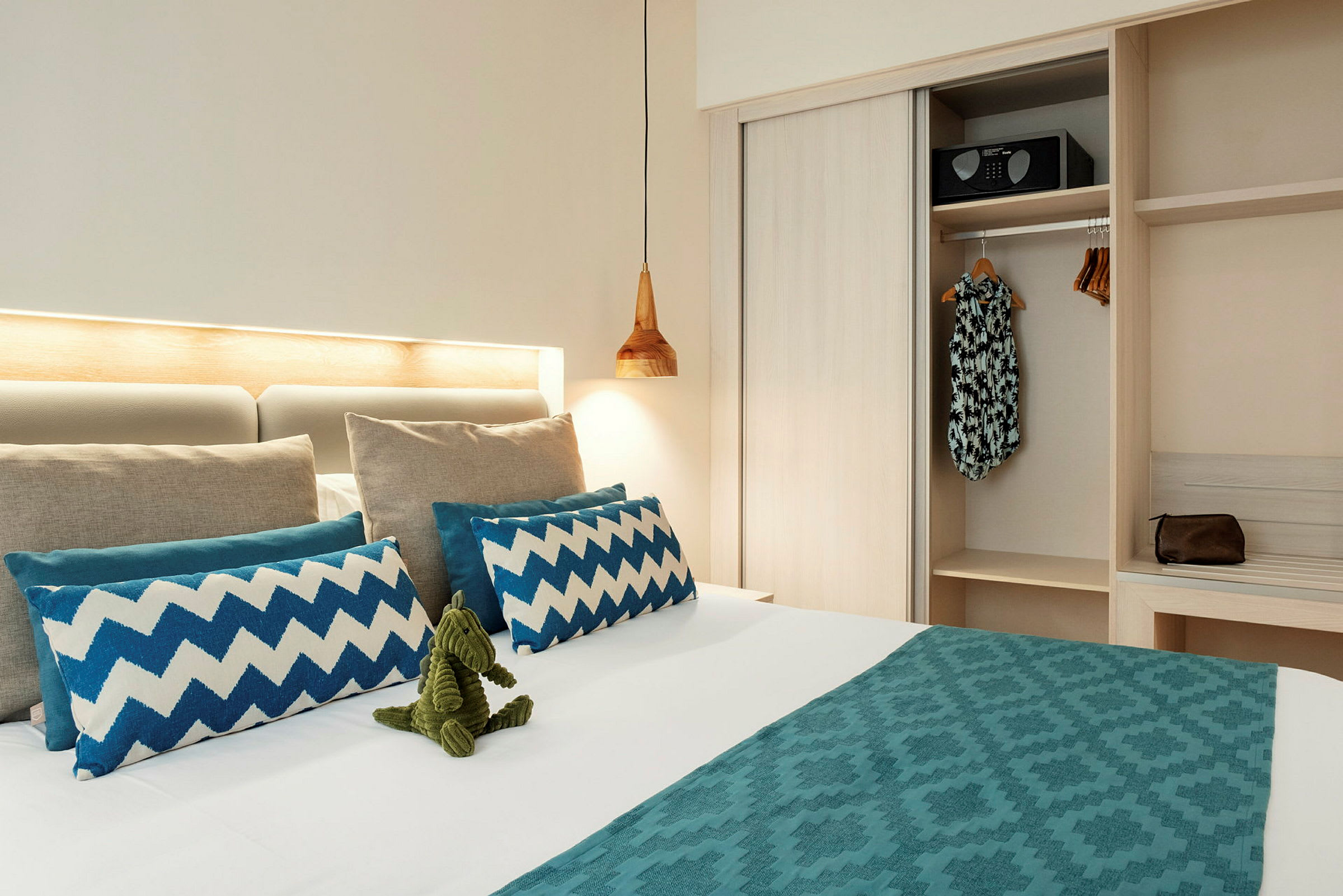 Ett hotellrum med en bäddad säng som har dekorativa blå kuddar och en grön leksak. En garderob med kläder och en säkerhetsbox är synlig bredvid. (AI-genererad text)
