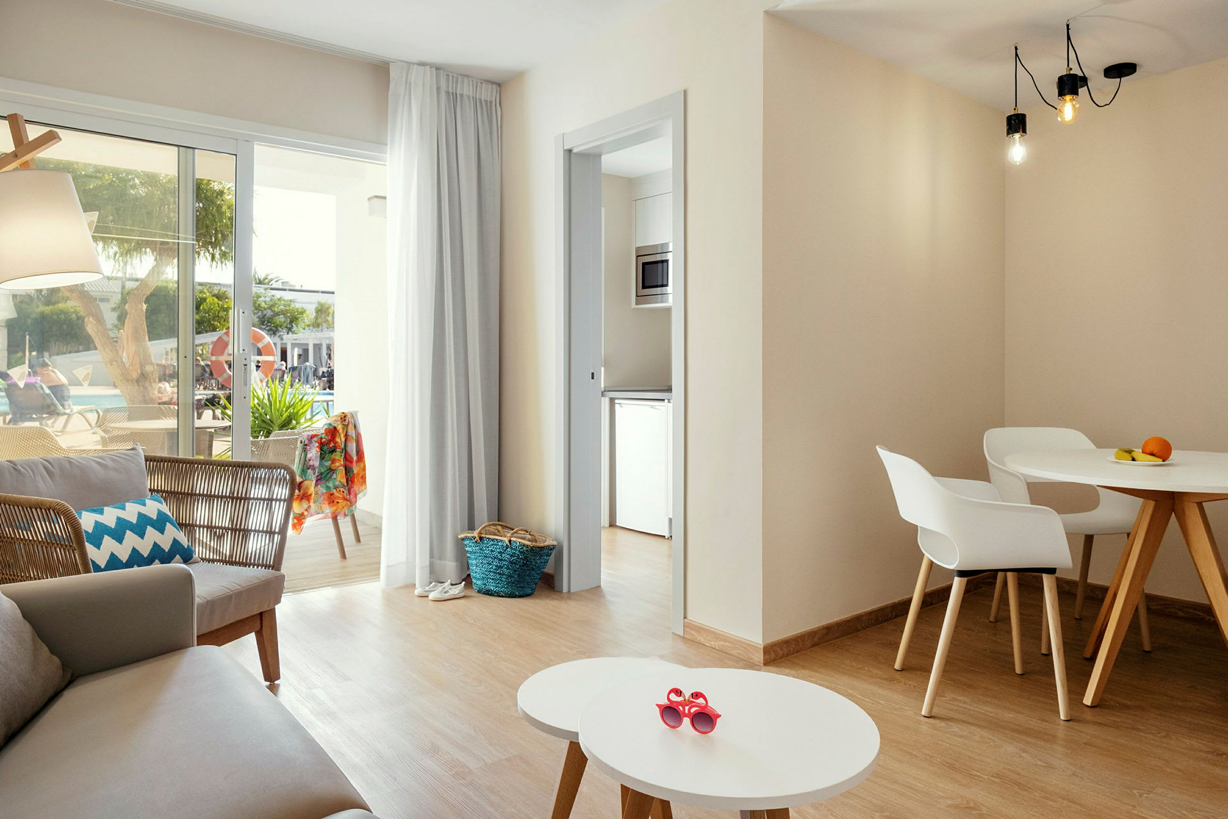 Ett ljust och modernt inrett vardagsrum med sittgrupp, matbord och stora fönster mot en terrass med utsikt över en pool. (AI-genererad text)