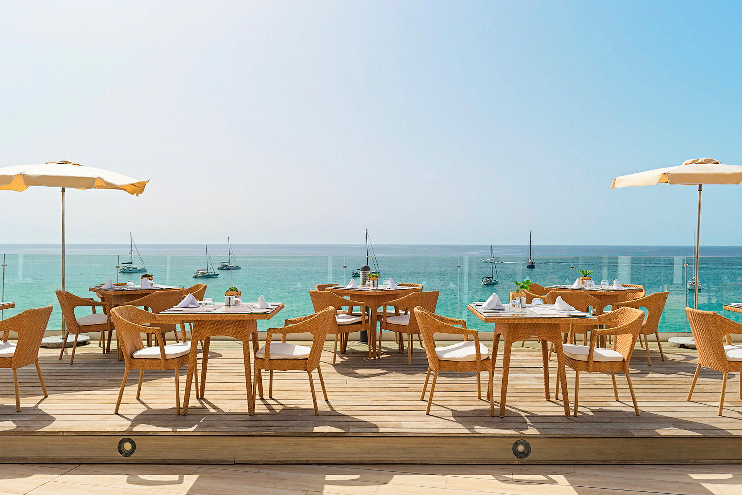 Utomhusrestaurang med utsikt över havet. Flera bord och stolar står på en terrass med parasoller och segelbåtar syns i bakgrunden. (AI-genererad text)