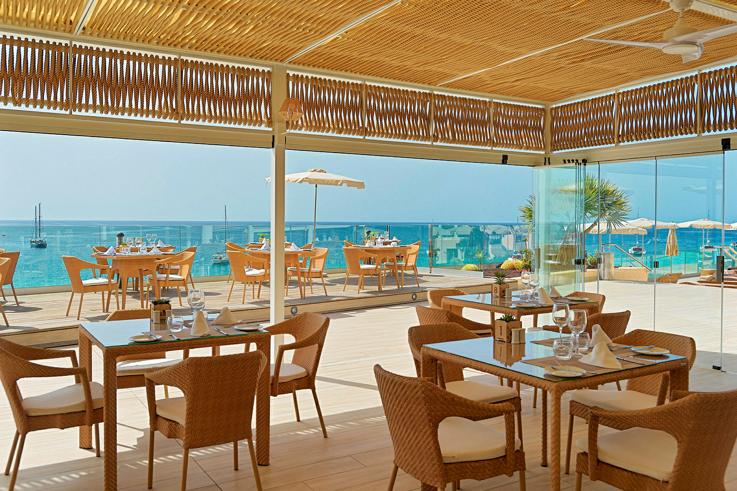 Bild av en restaurang med flätade stolar och glasbord, belägen vid havet med utsikt över en blå vattenhorisont. (AI-genererad text)