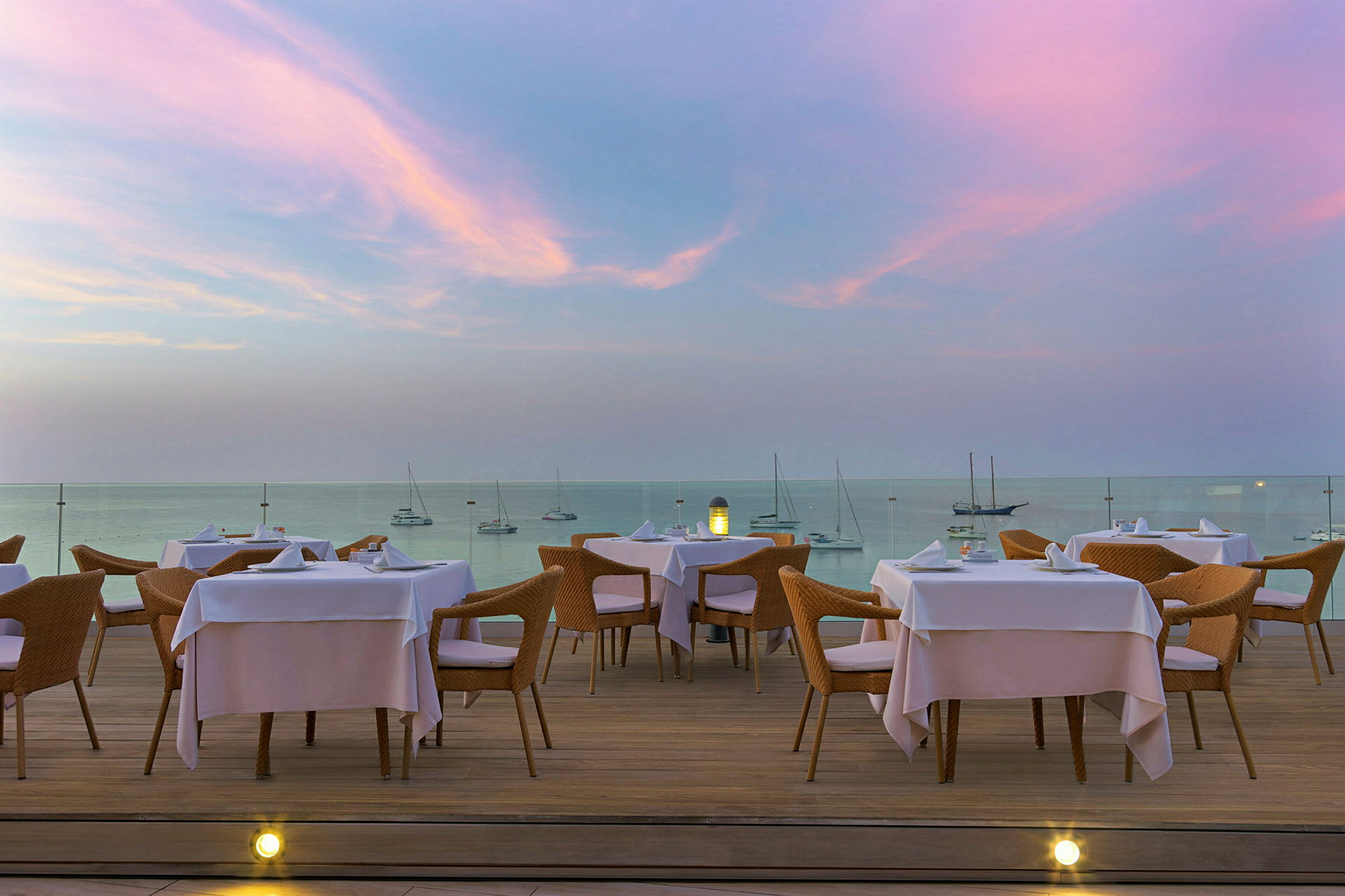 En restaurangterrass med bord och stolar, med utsikt över havet under en rosa och blå kvällshimmel. (AI-genererad text)