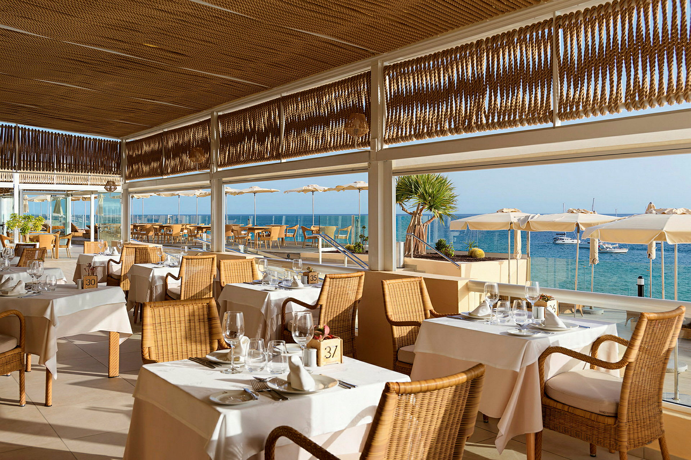 Restaurang med rottingstolar och dukade bord, belägen vid havet med utsikt över vattnet och båtar. (AI-genererad text)