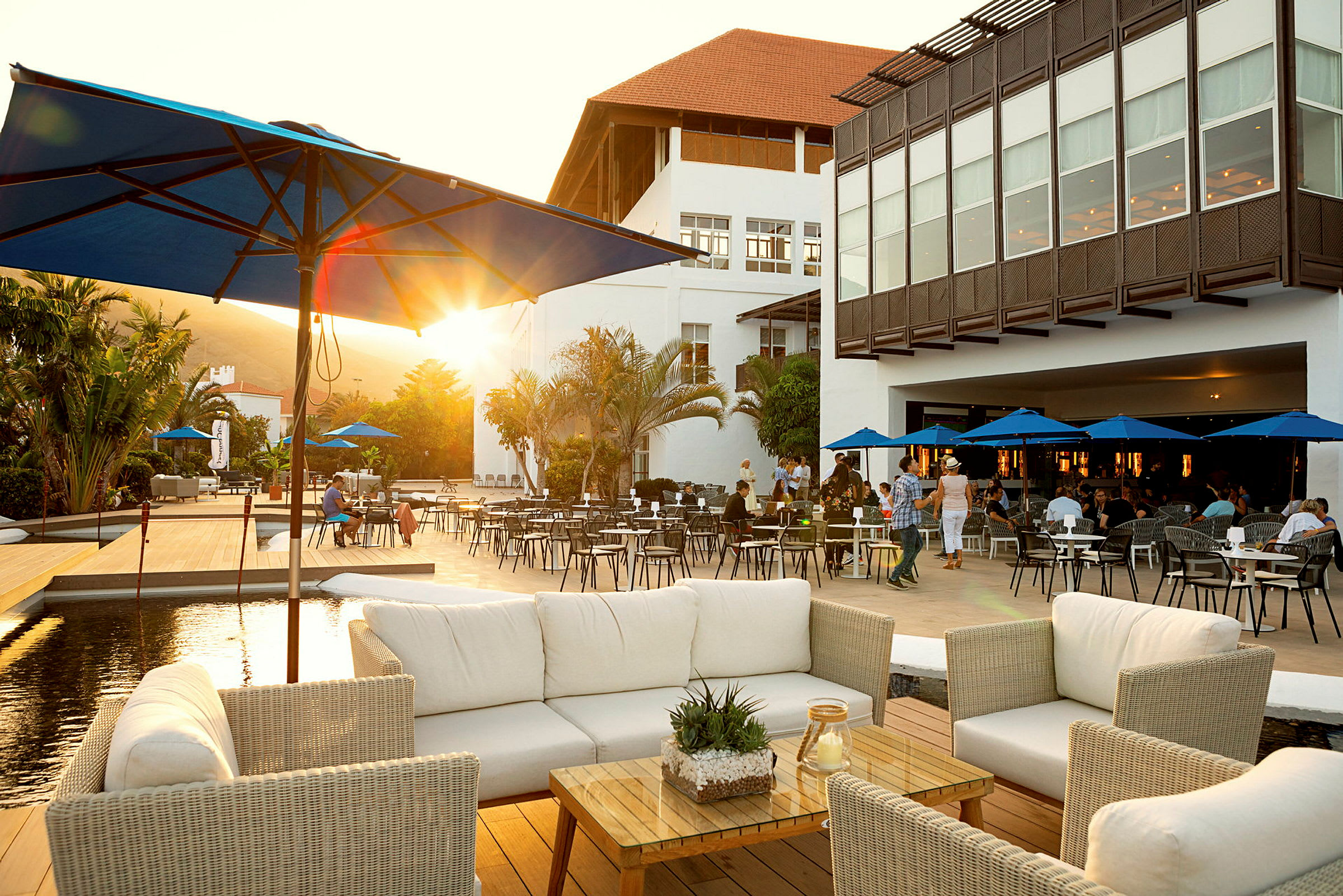 En restaurangterrass med vita rottingmöbler och parasoller, omgiven av ett palmbeklätt område och ett stort vitt byggnadskomplex i bakgrunden. (AI-genererad text)