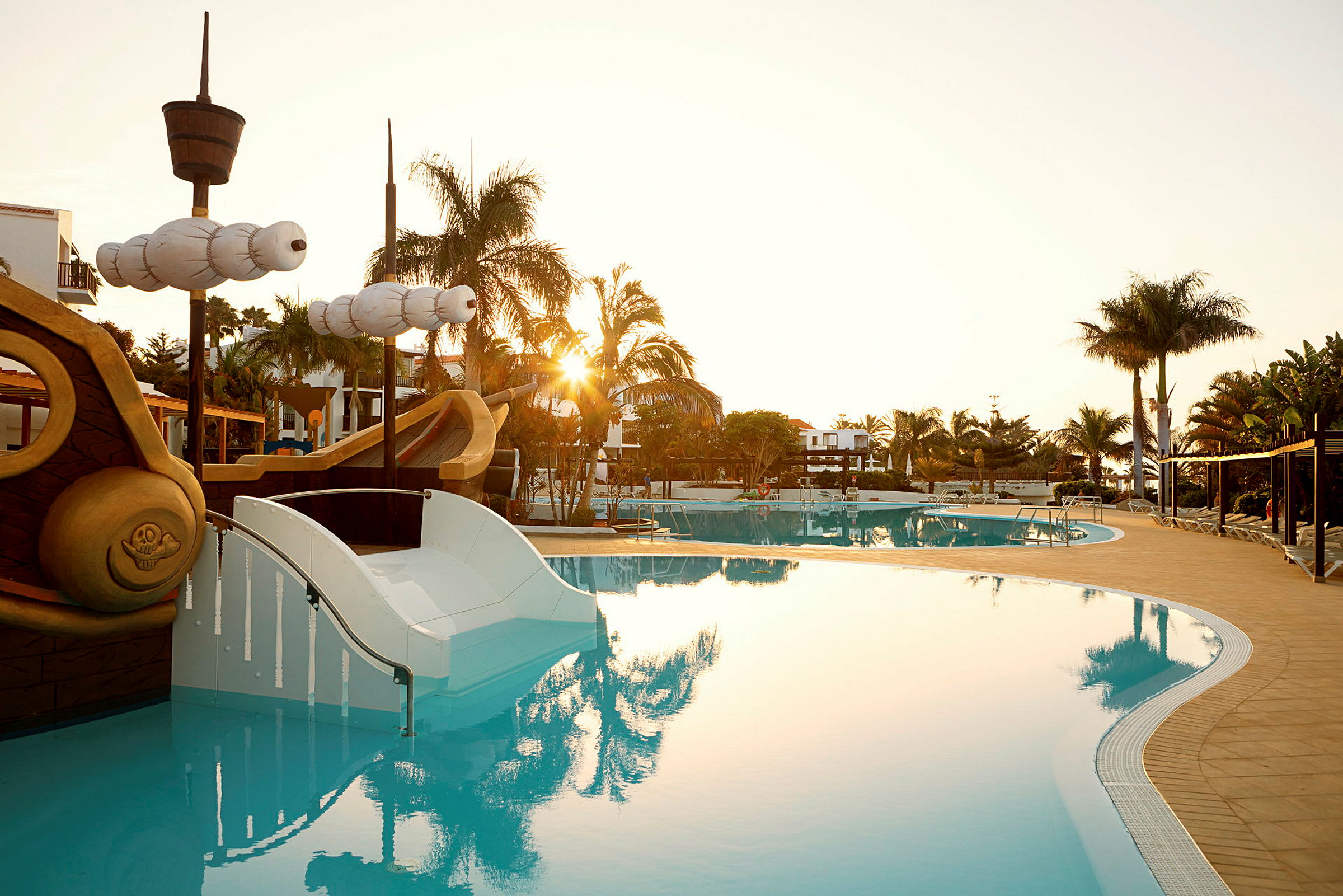 En pool vid ett semesterområde med en solnedgång i bakgrunden. Det finns lekfulla element som en åkattraktion formad som ett piratskepp och palmer runtom. (AI-genererad text)