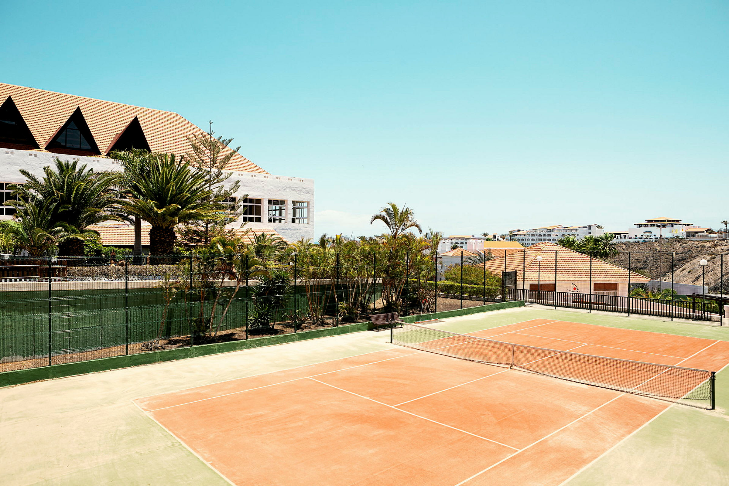 En tennisbana i solen med omgivande grönska och byggnader i bakgrunden. (AI-genererad text)