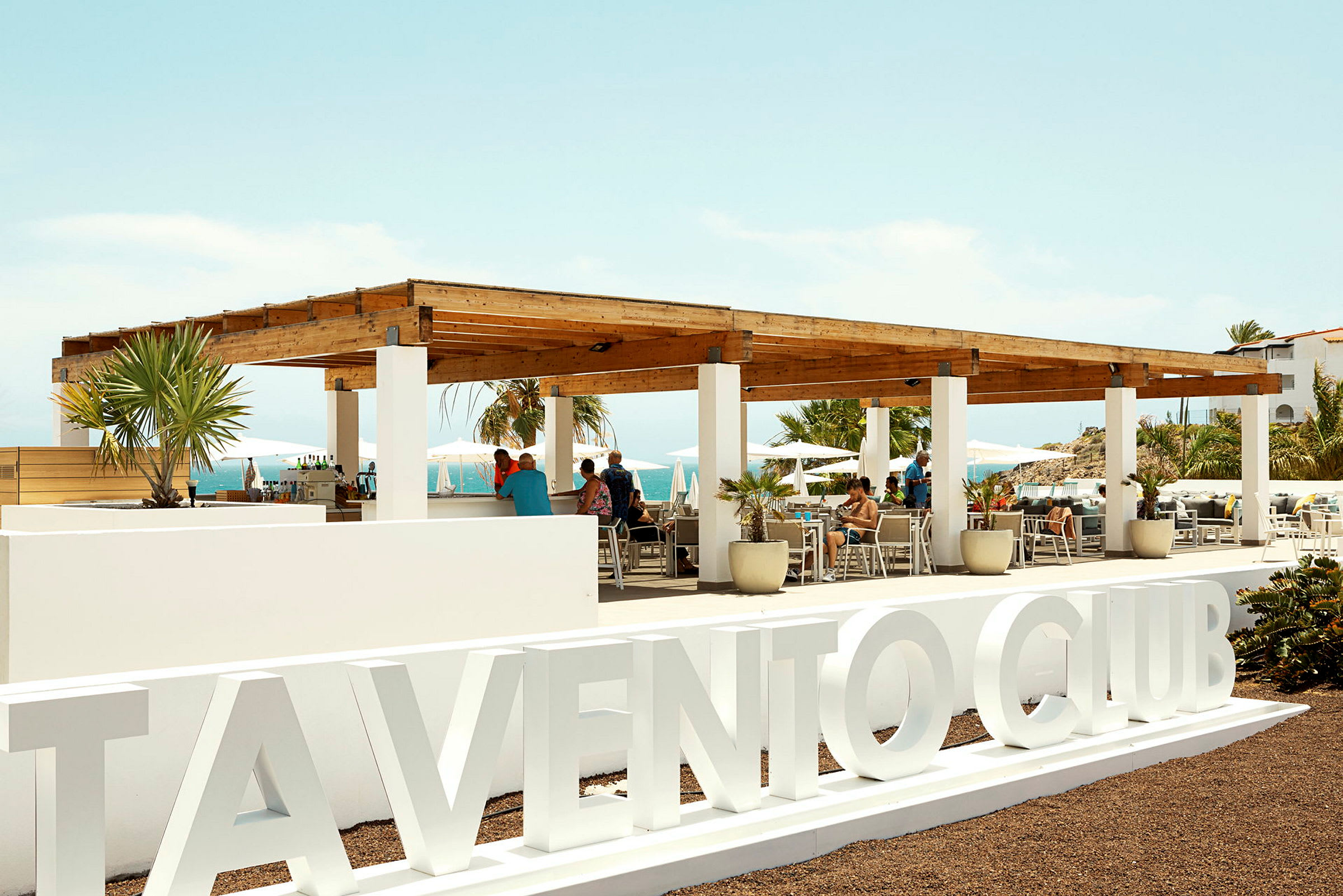 En utomhusbar och sittplats med en träpergola och vita stolpar. Skylten 'Vento Club' syns i förgrunden, och människor sitter och njuter av utsikten. (AI-genererad text)