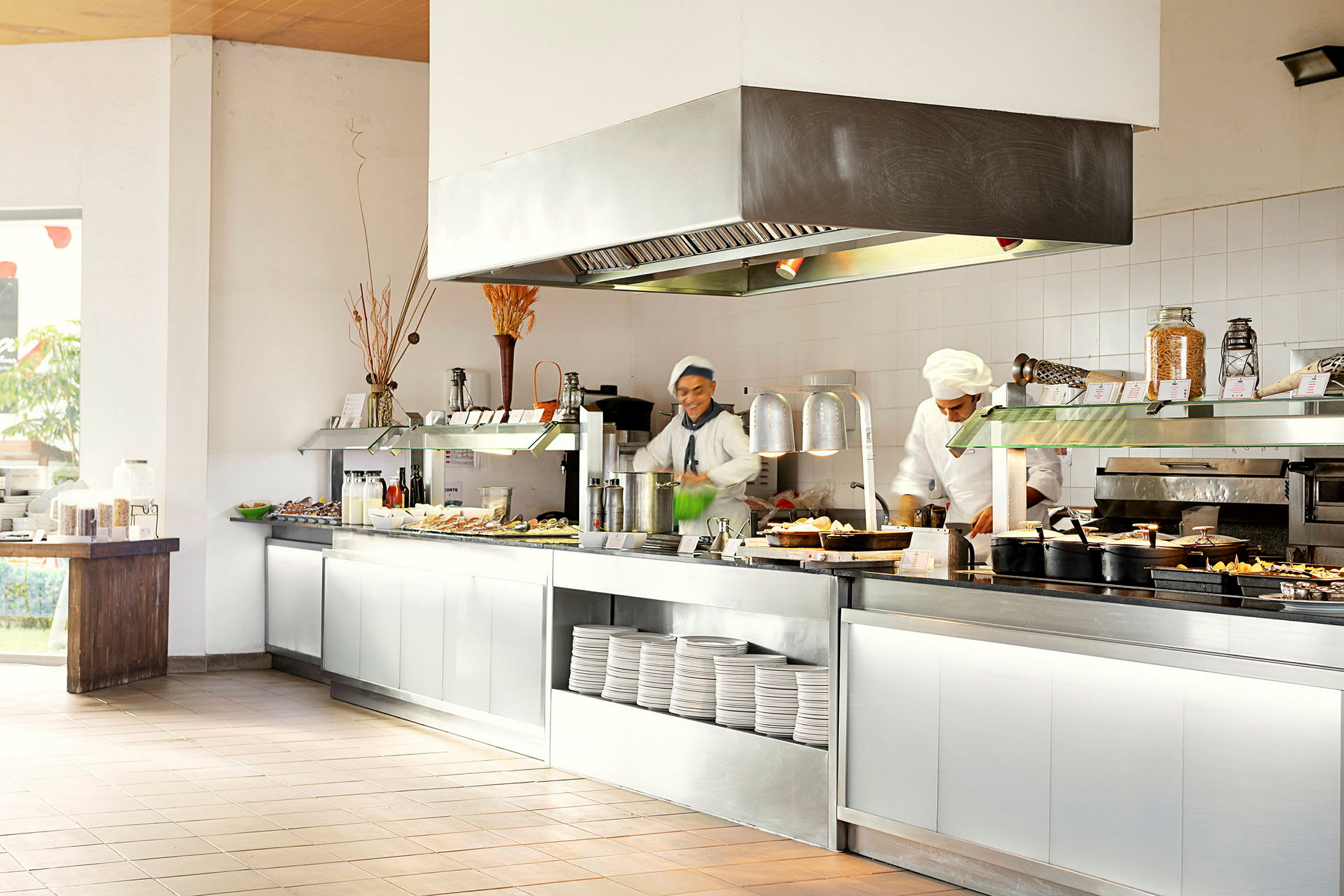 En restaurangbuffé med olika maträtter och tallrikar upplagda på ett serveringsbord. Kockar förbereder mat. (AI-genererad text)