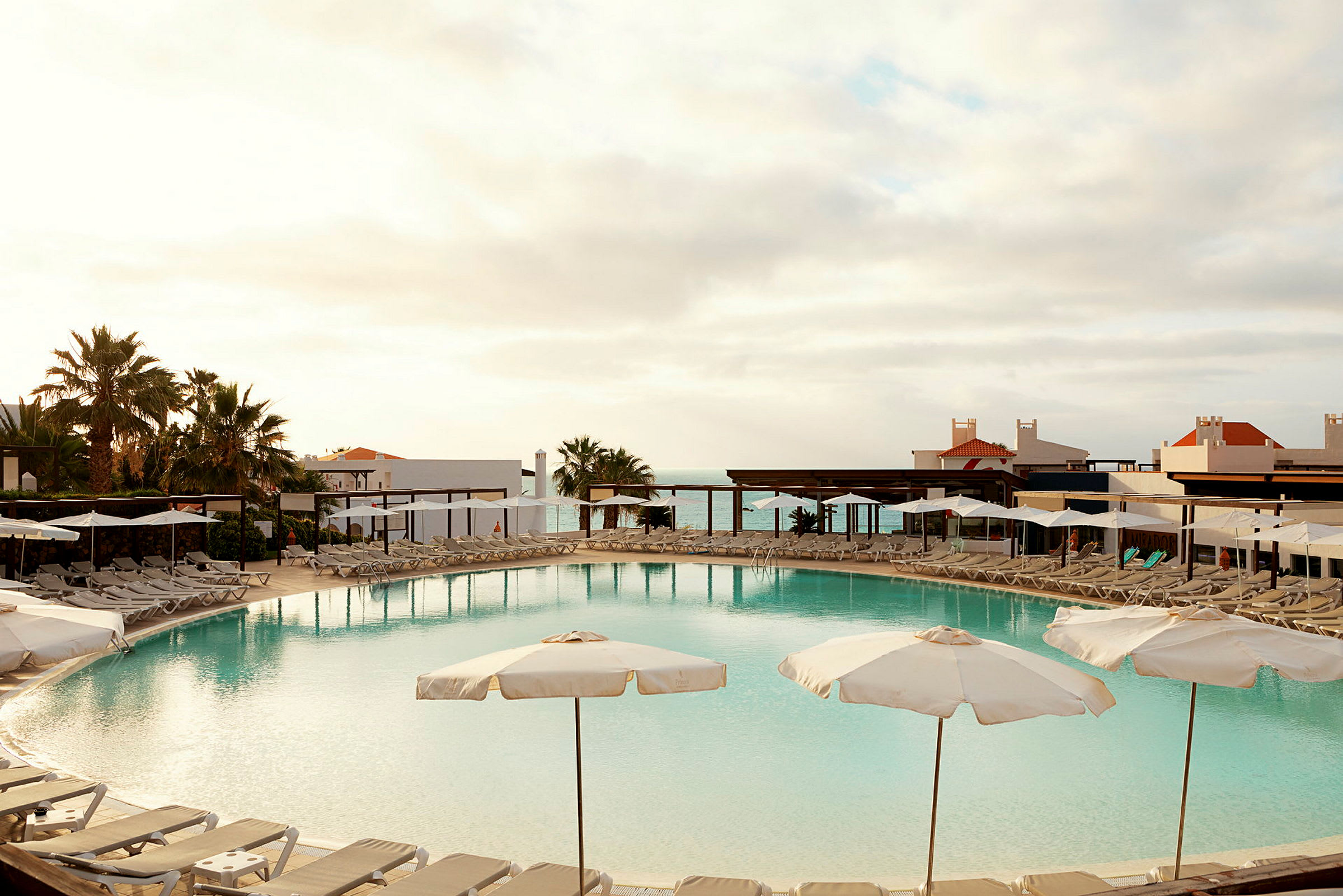 En pool omgiven av solstolar och parasoller med en utsikt över havet i bakgrunden. (AI-genererad text)