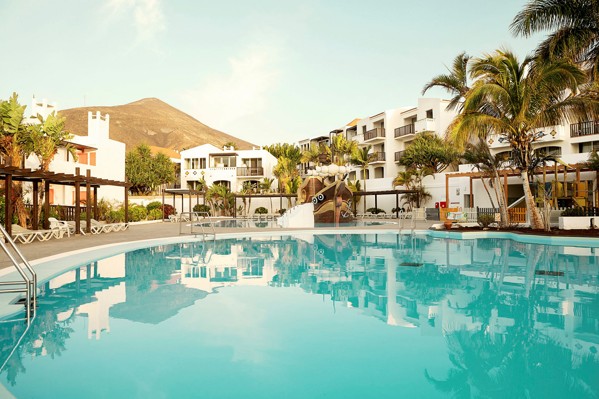 En bild på en pool framför vita hotellbyggnader, med palmer och ett berg i bakgrunden. (AI-genererad text)